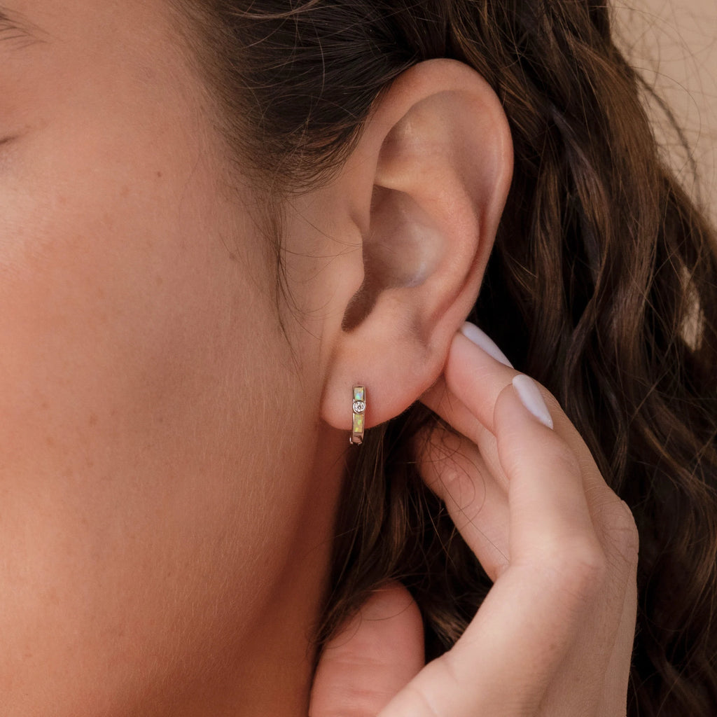 A hand touches a small Opal Inlay Diamond Huggie earring worn on a woman with wavy brown hair, highlighting the earring’s elegant, subtle shine.