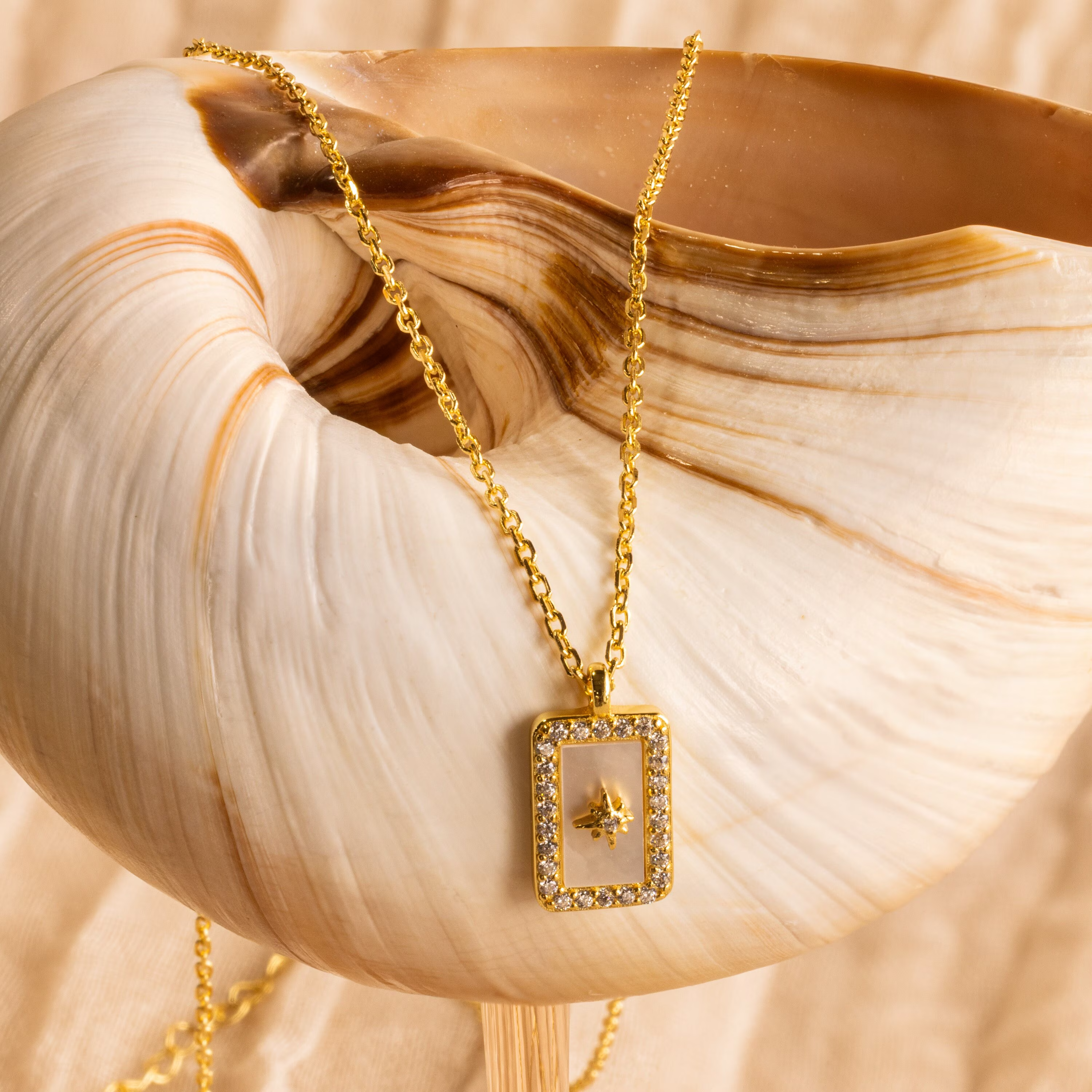The Pave Pearl North Star Necklace features a gold rectangular pendant adorned with small crystals, elegantly displayed on a large brown-marked seashell—perfect for fans of celestial jewelry.