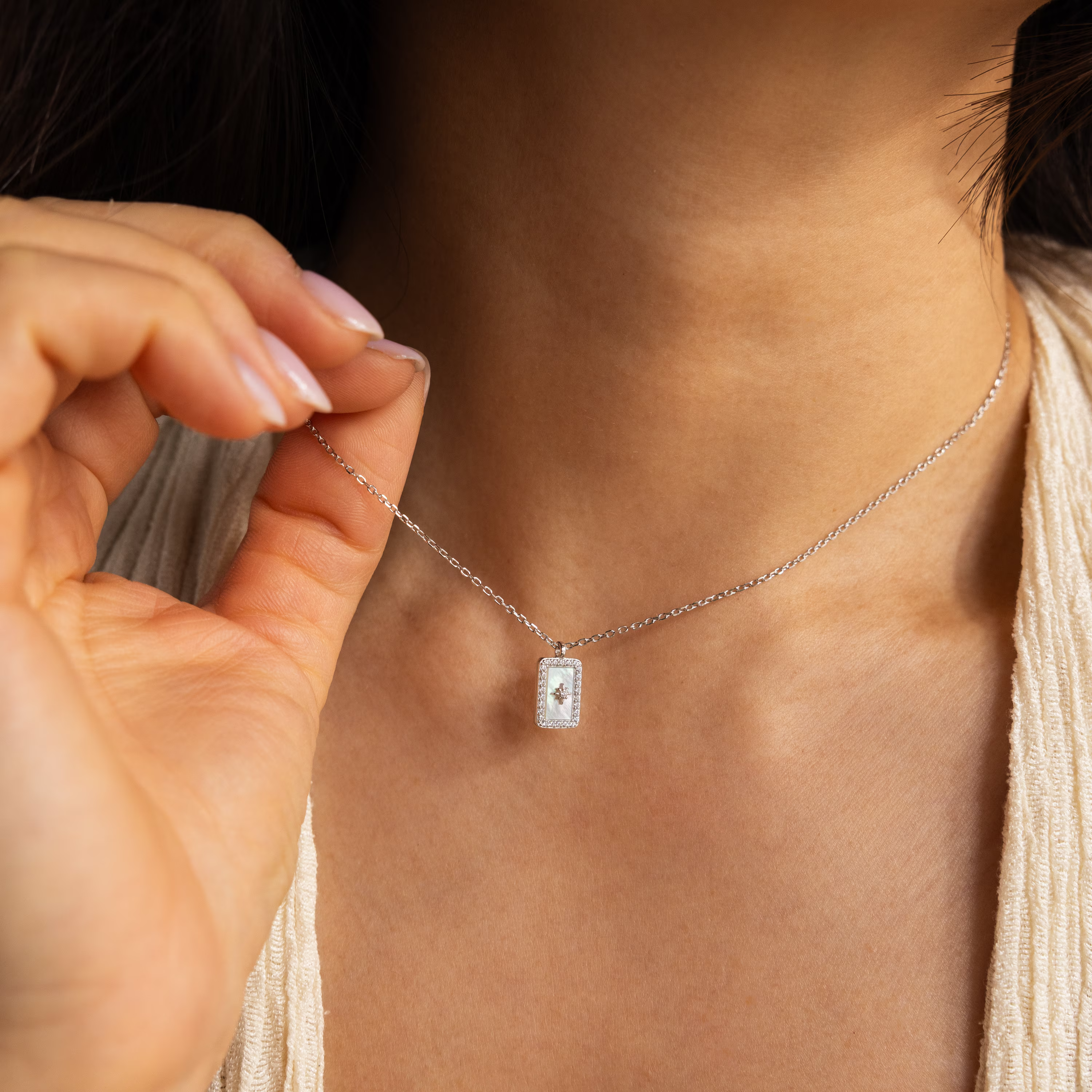 A woman holds the Pave Pearl North Star Necklace with its rectangular pendant near her neck, highlighting the celestial elegance of this jewelry piece.