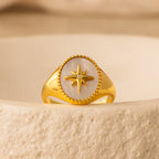 The North Star Signet Ring features an eight-pointed star and a small gemstone, displayed on a textured beige surface—a striking statement piece of jewelry.