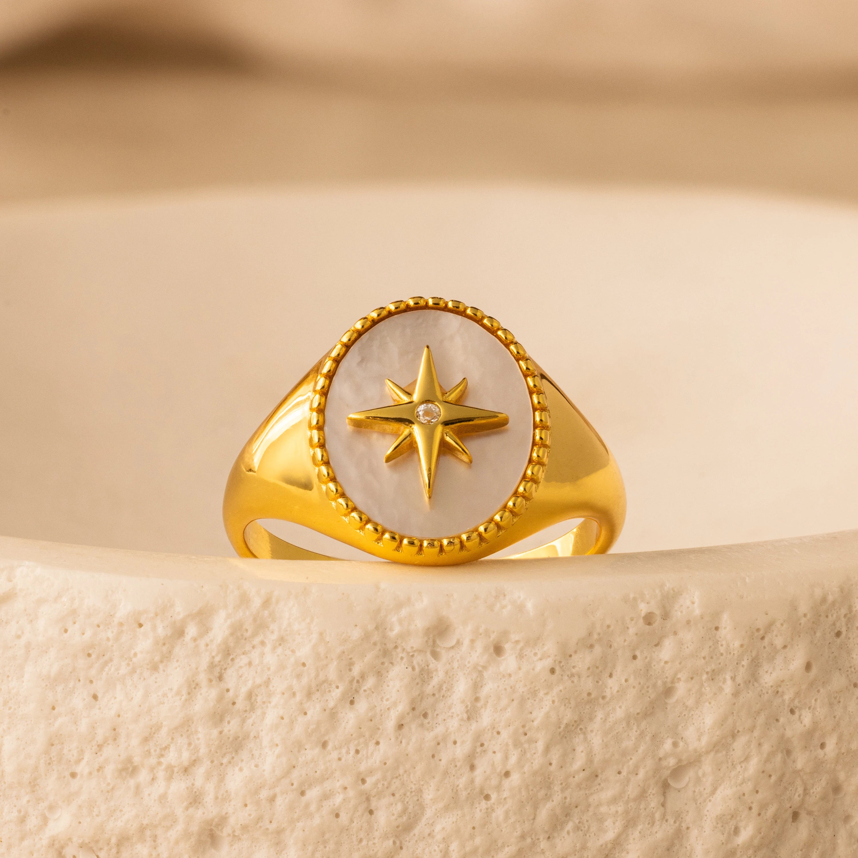 The North Star Signet Ring features an eight-pointed star and a small gemstone, displayed on a textured beige surface—a striking statement piece of jewelry.