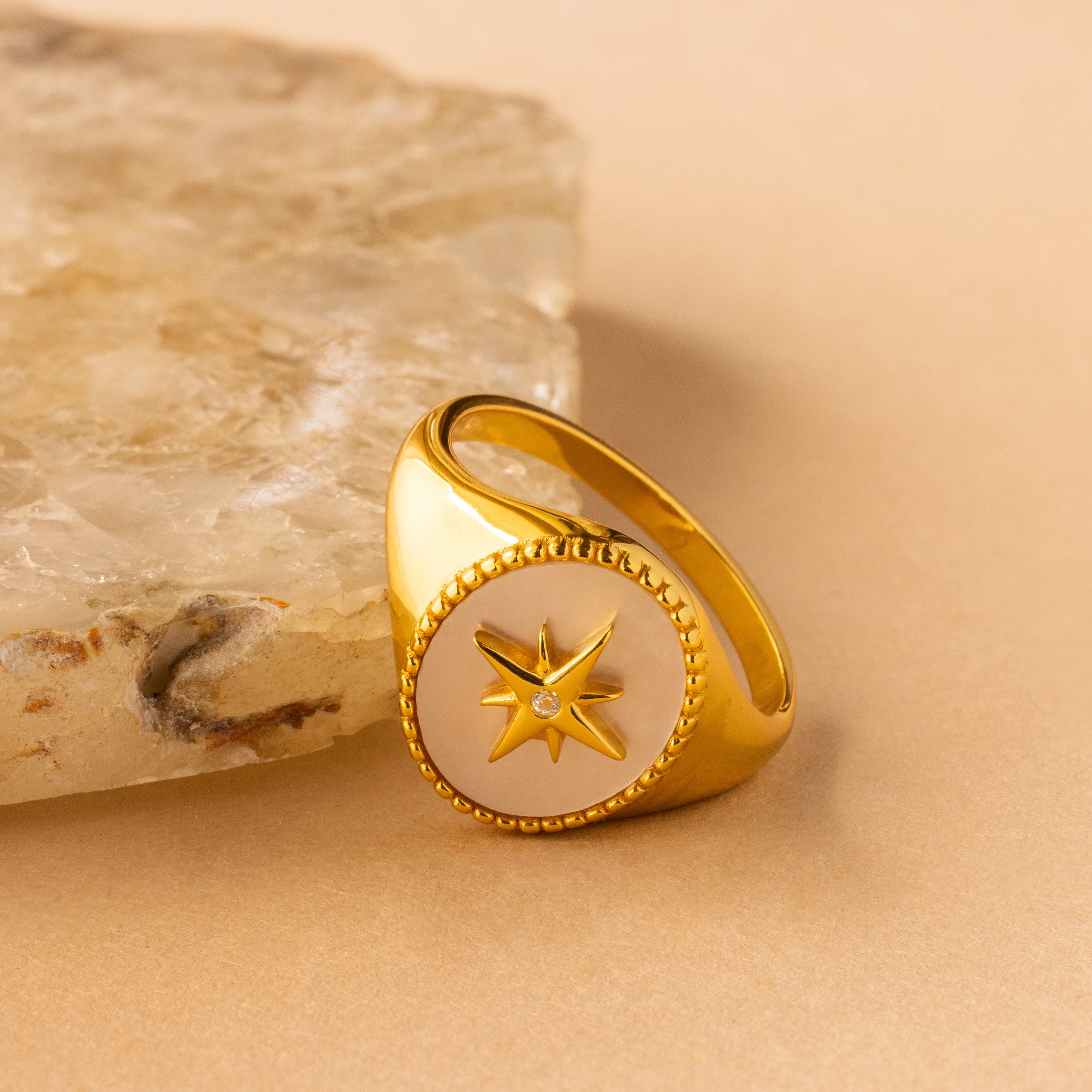 The North Star Signet Ring features a star design on a white circular face, showcased beside a textured stone on a beige surface—a standout statement piece ideal for any jewelry collection.