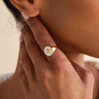 A hand touches the face, highlighting the North Star Signet Ring—a striking gold statement piece featuring a white oval with a star design.