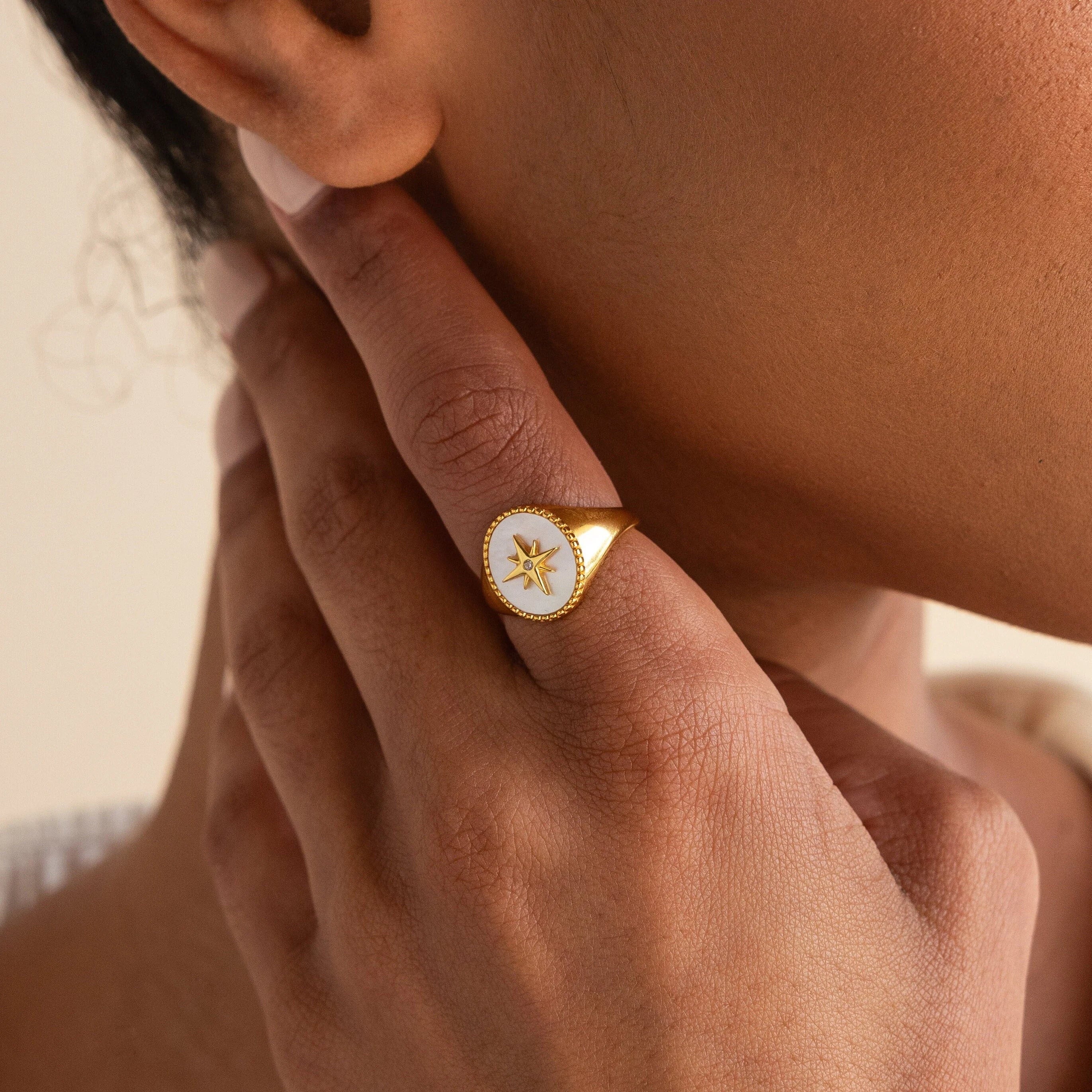A hand touches the face, highlighting the North Star Signet Ring—a striking gold statement piece featuring a white oval with a star design.