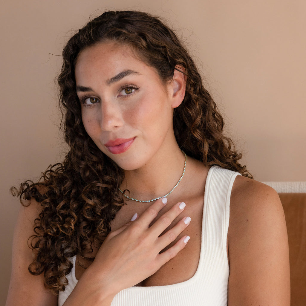 Woman with curly brown hair, wearing a white top and the Aquamarine Tennis Choker Necklace, touches her chest and smiles softly.