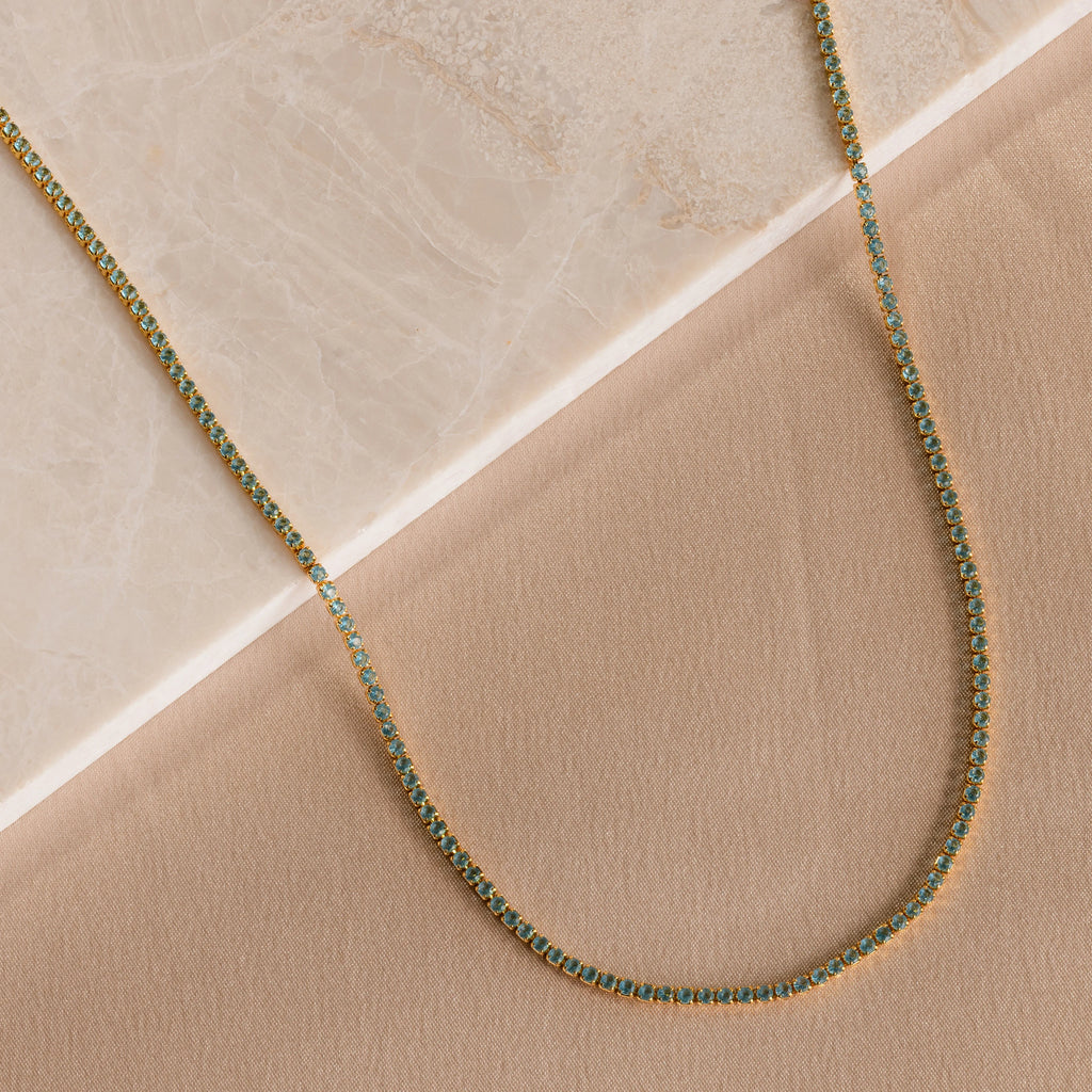 The Aquamarine Tennis Choker Necklace features a delicate gold chain with aquamarine stones, beautifully displayed on beige marble and fabric—an elegant, understated choice for bridal or wedding jewelry.