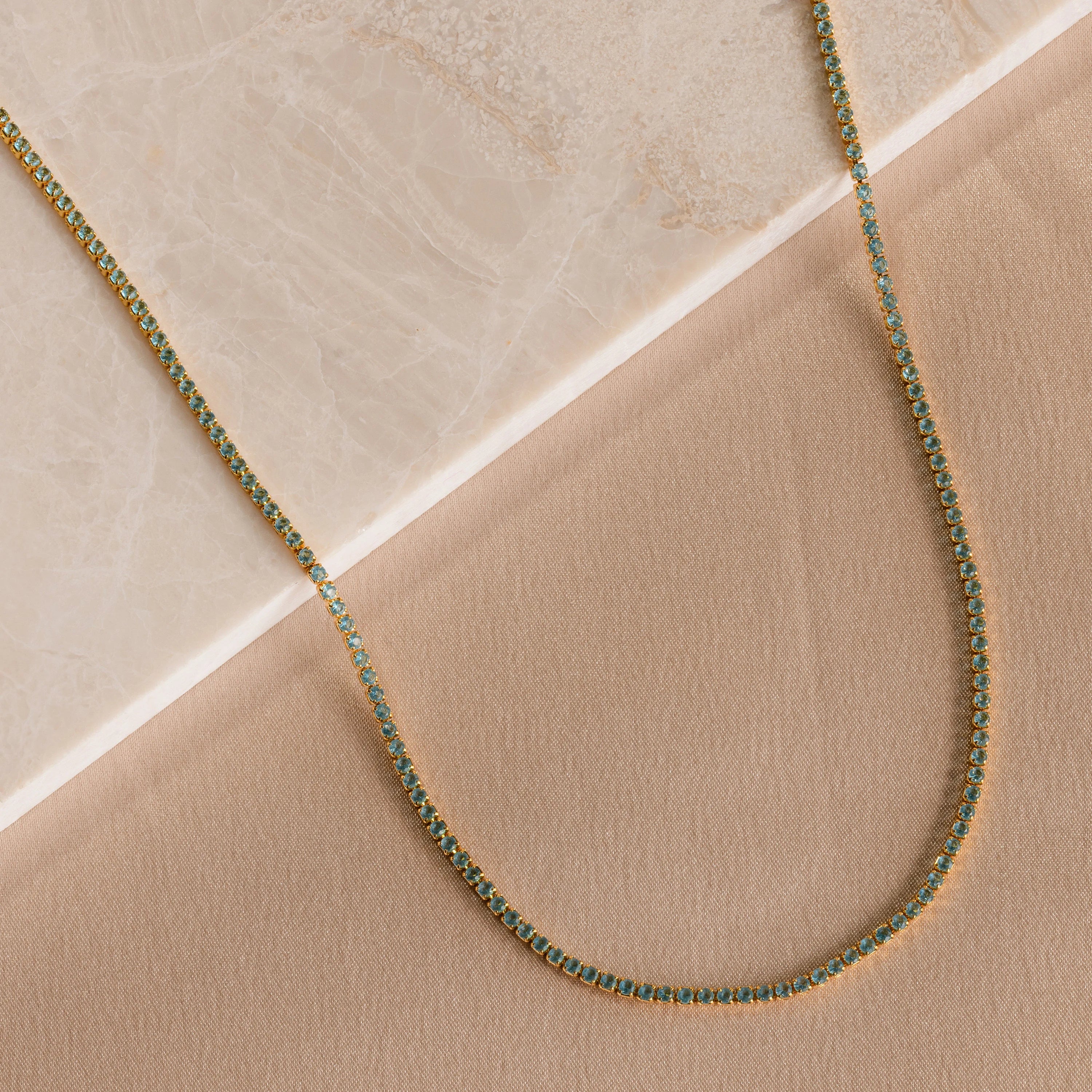 The Aquamarine Tennis Choker Necklace features a delicate gold chain with aquamarine stones, beautifully displayed on beige marble and fabric—an elegant, understated choice for bridal or wedding jewelry.