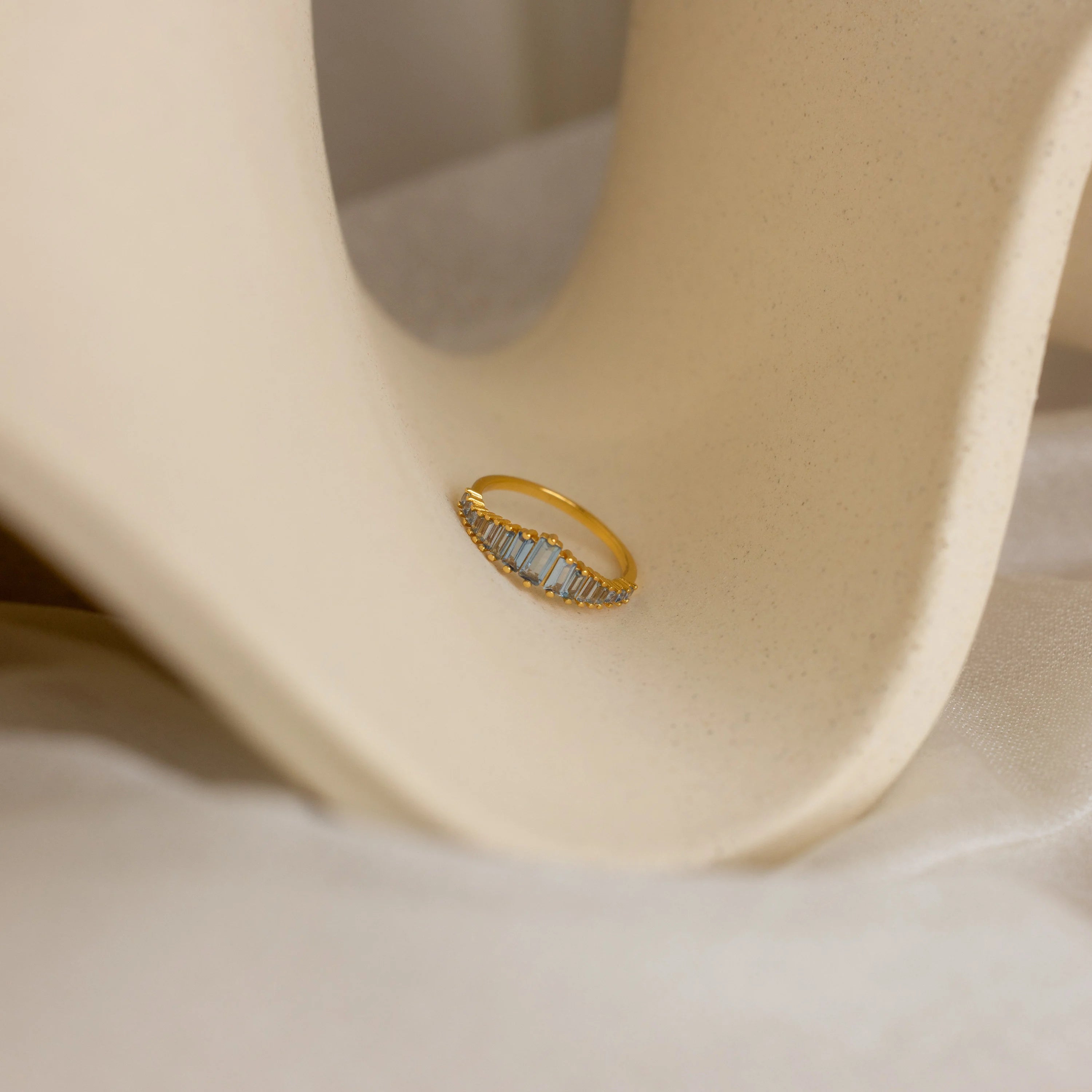 The Art Deco Aquamarine Ring, featuring rectangular clear stones, rests on a smooth curved beige surface, capturing vintage glamour.