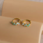 The Ombre Opal Huggies are gold hoop earrings featuring small oval opal stones, displayed on a beige curved surface and glowing beautifully in the light.
