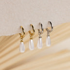 Two pairs of Pearl Teardrop Huggies—one gold, one silver—are shown on textured paper. These hoop earrings are ideal for brides or a chic beach wedding style.