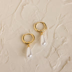 Pearl Teardrop Huggies: Gold hoop earrings with dangling teardrop-shaped pearls on a textured cream background, ideal for beach weddings or as elegant accessories for your special day.