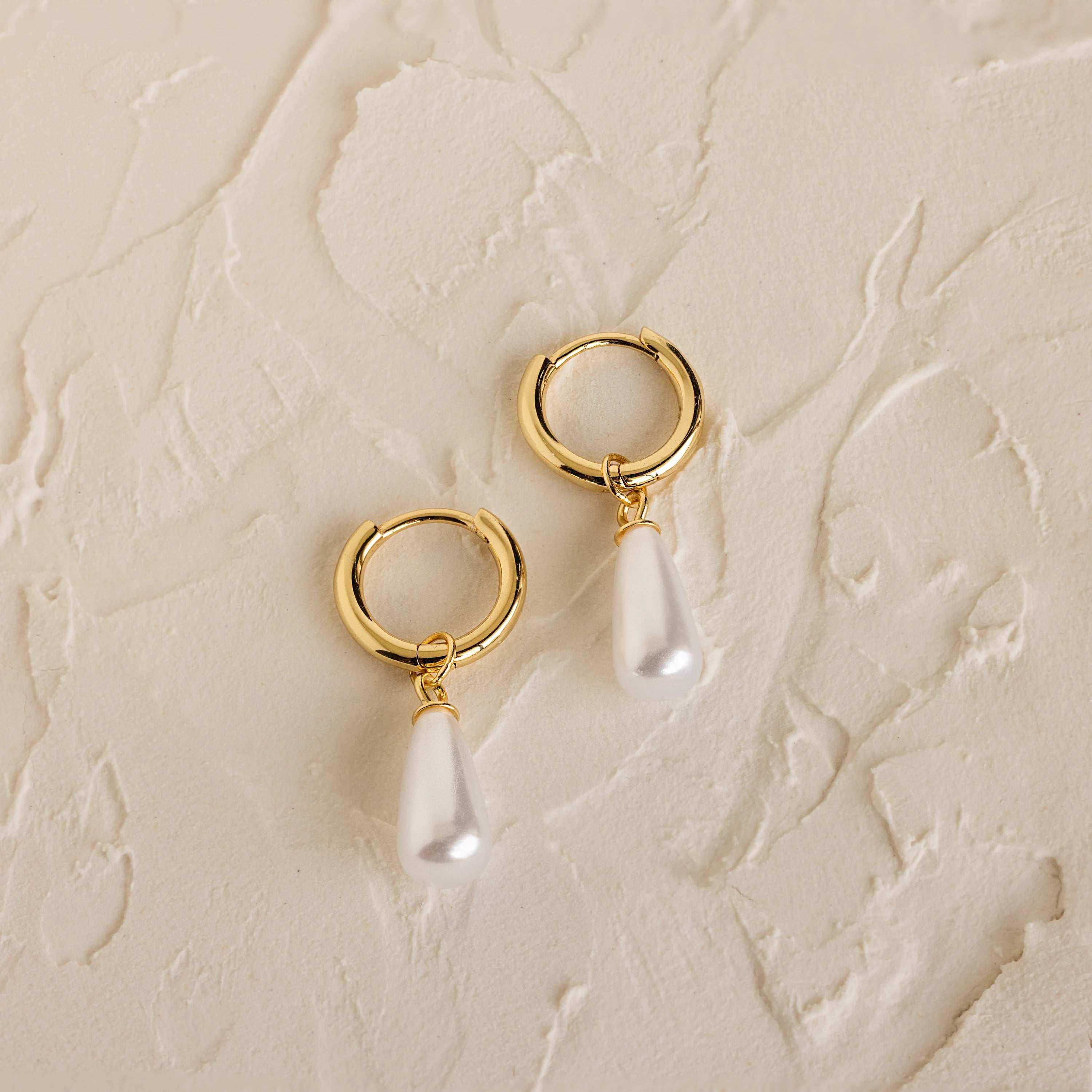 Pearl Teardrop Huggies: Gold hoop earrings with dangling teardrop-shaped pearls on a textured cream background, ideal for beach weddings or as elegant accessories for your special day.