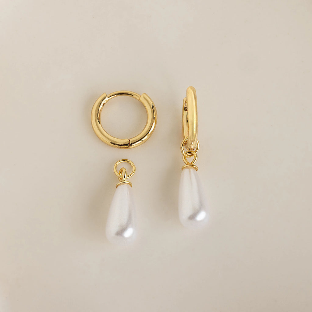 The Pearl Teardrop Huggies feature two gold hoops—one plain, one with a teardrop-shaped white pearl charm—perfect as elegant bridal earrings or for a beach wedding. Shown on a cream background.