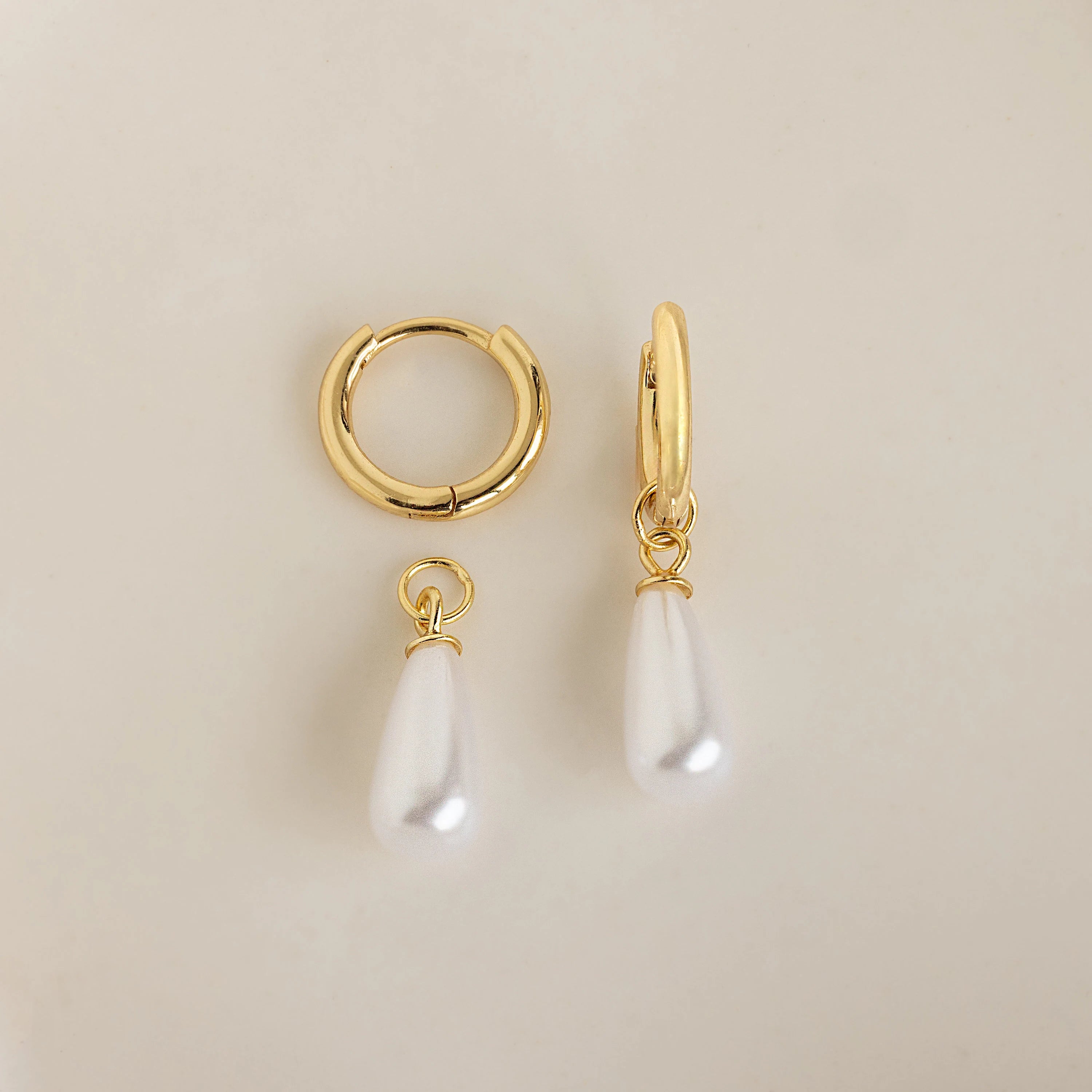 The Pearl Teardrop Huggies feature two gold hoops—one plain, one with a teardrop-shaped white pearl charm—perfect as elegant bridal earrings or for a beach wedding. Shown on a cream background.