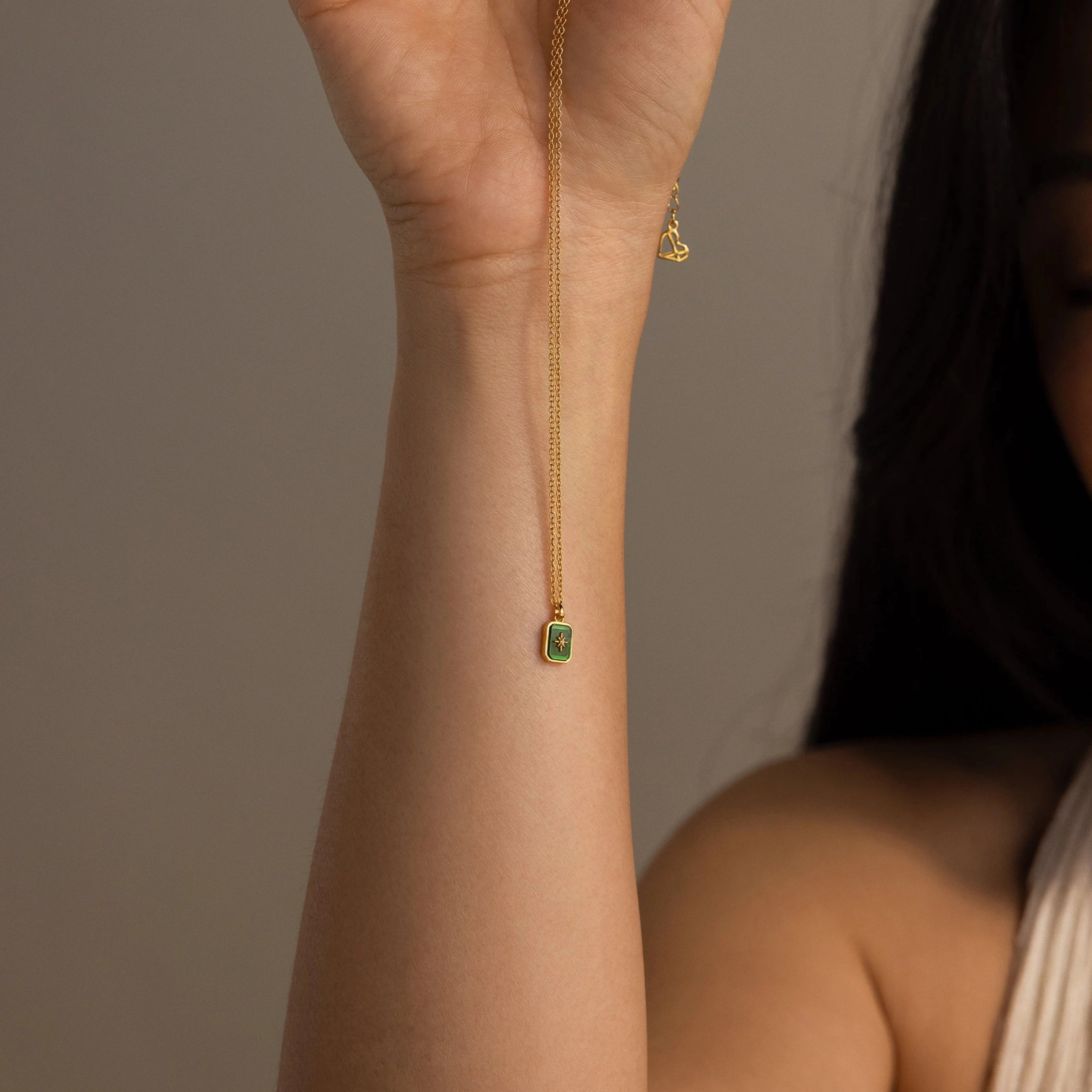 Someone displays the North Star Birthstone Necklace, featuring a gold finish and a small green crystal pendant, against a neutral background.