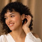 A smiling woman with curly hair wears Pearl Teardrop Huggies and a white top, touching her earrings in front of a beige curtain.