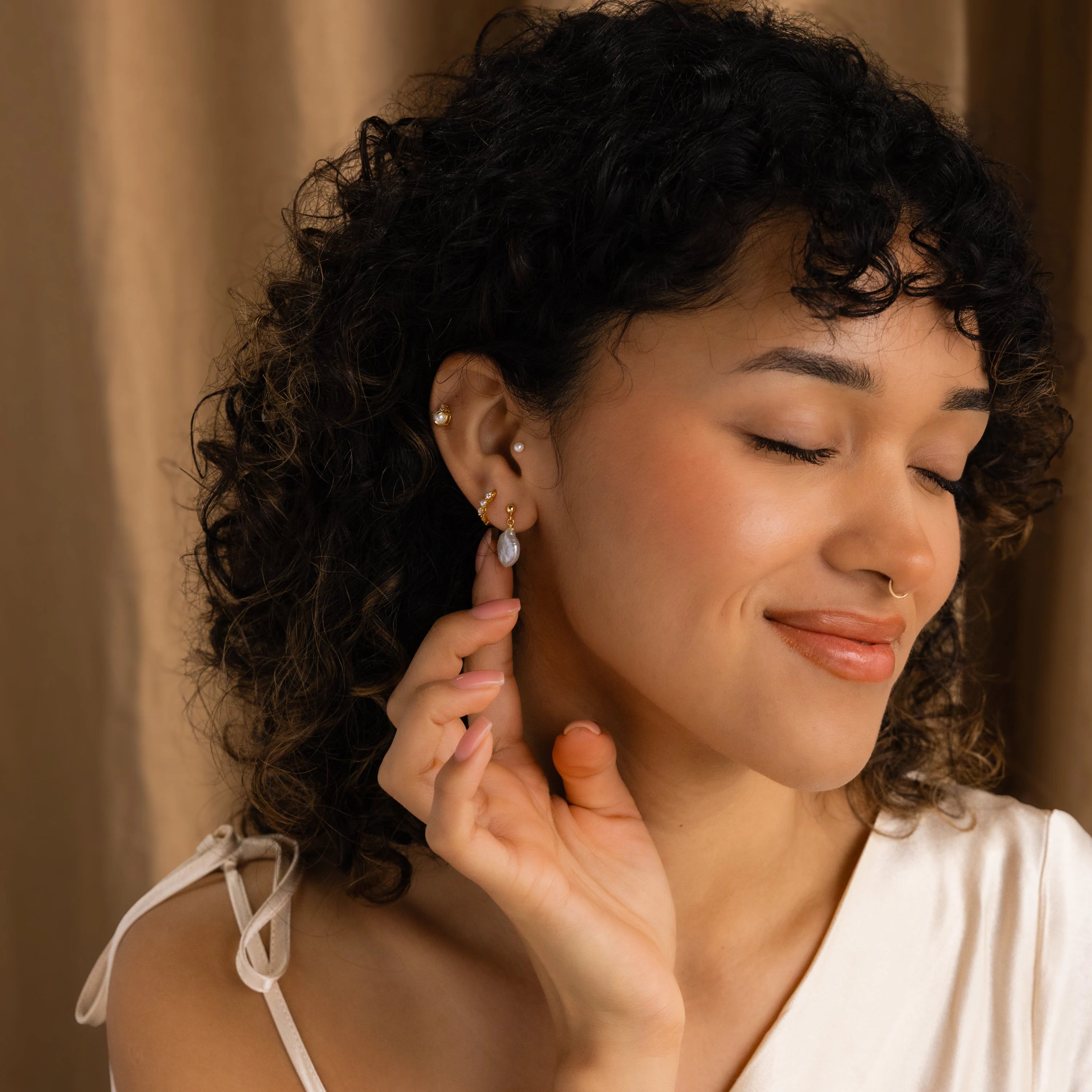A woman with curly hair smiles with eyes closed, touching her Round Pearl Drop Earrings while wearing a white top against a beige background.