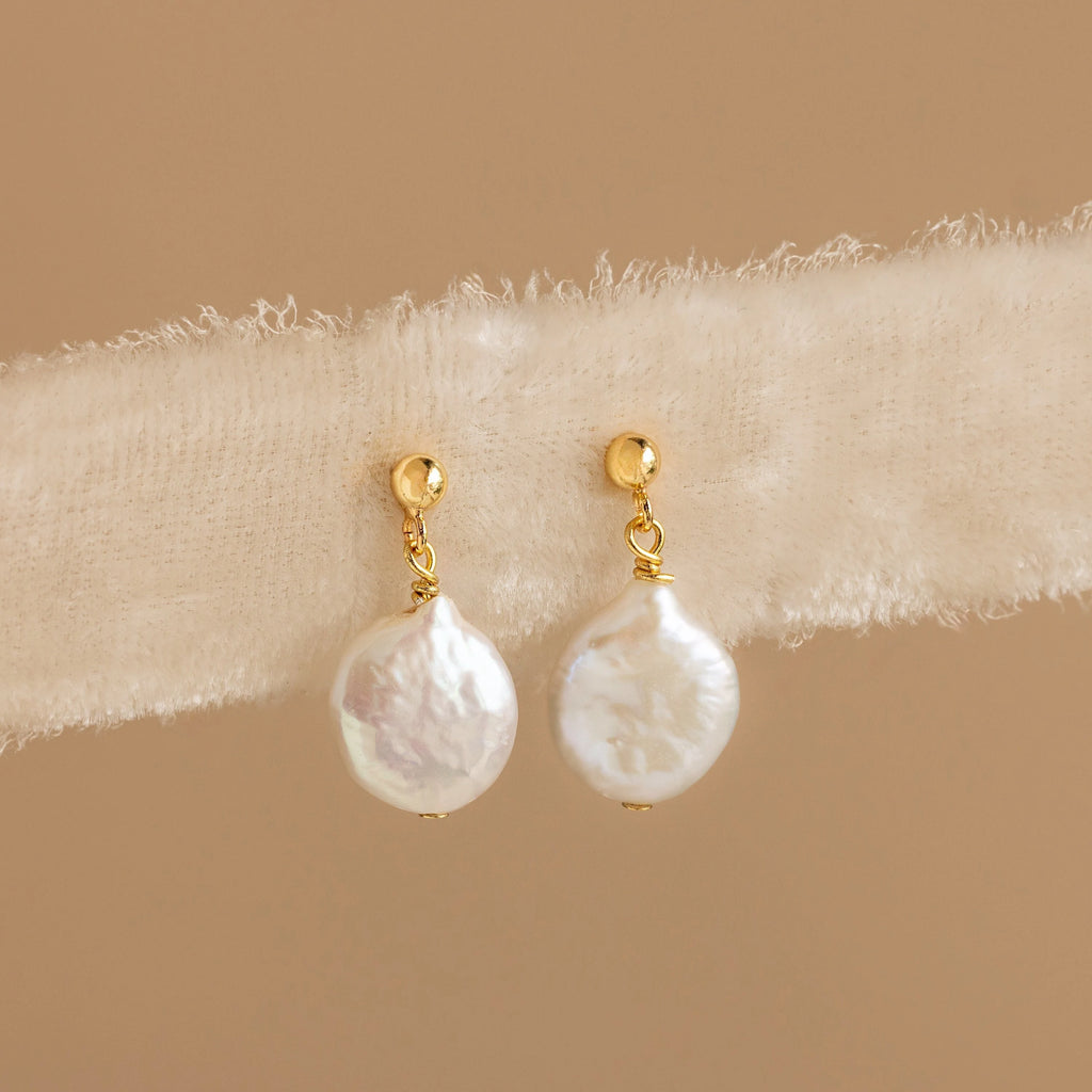 Round Pearl Drop Earrings with gold accents drape elegantly on a soft, beige textured fabric background.