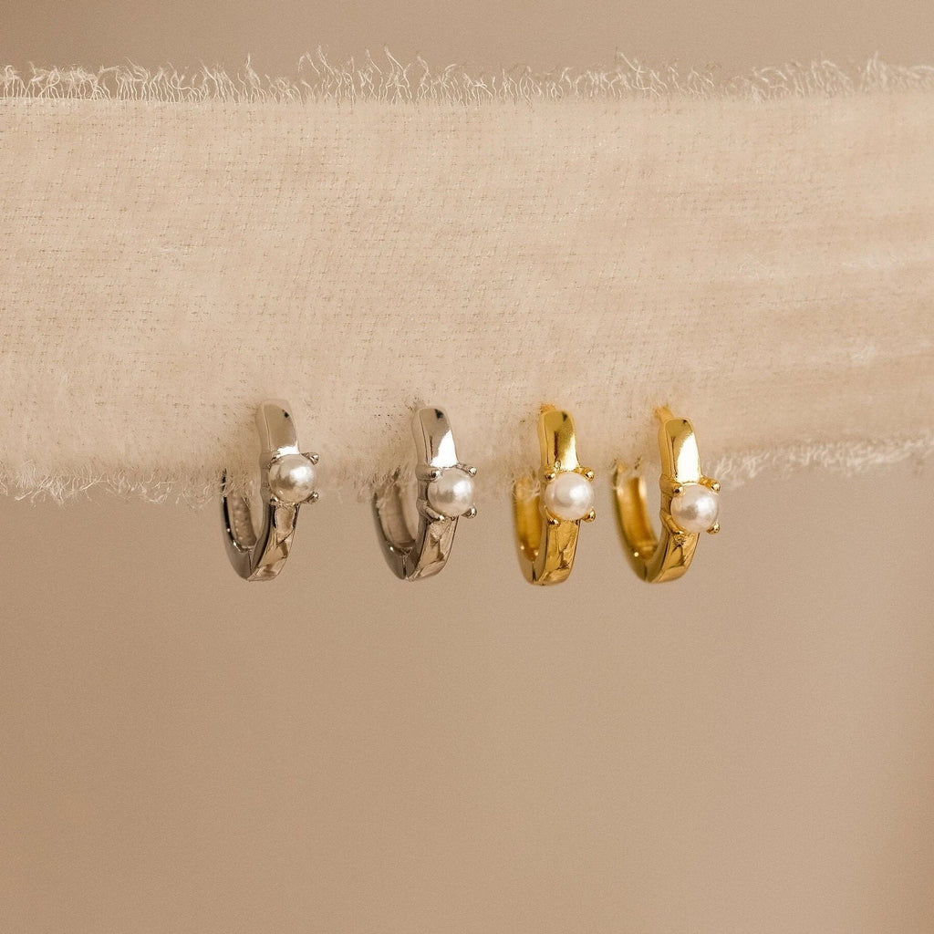 The Tiny Pearl Huggies—one silver, one gold—feature classic pearls on modern huggie hoops, elegantly displayed on textured fabric.