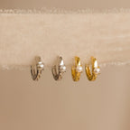 The Tiny Pearl Huggies—one silver, one gold—feature classic pearls on modern huggie hoops, elegantly displayed on textured fabric.