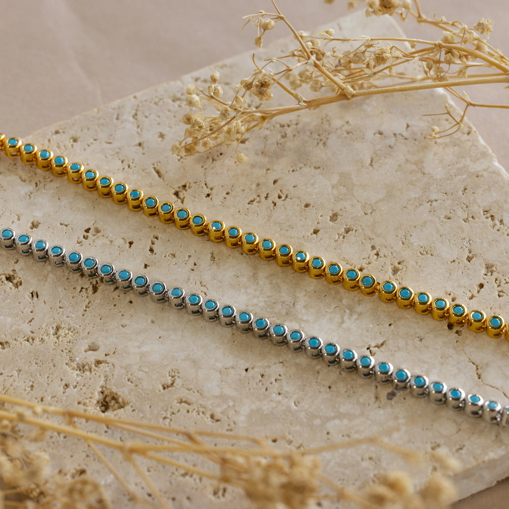 A gold and a silver beaded bracelet with turquoise stones rest on a textured stone surface beside dried flowers—perfect for pairing with the Turquoise Bezel Tennis Necklace.