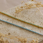 A gold and a silver beaded bracelet with turquoise stones rest on a textured stone surface beside dried flowers—perfect for pairing with the Turquoise Bezel Tennis Necklace.