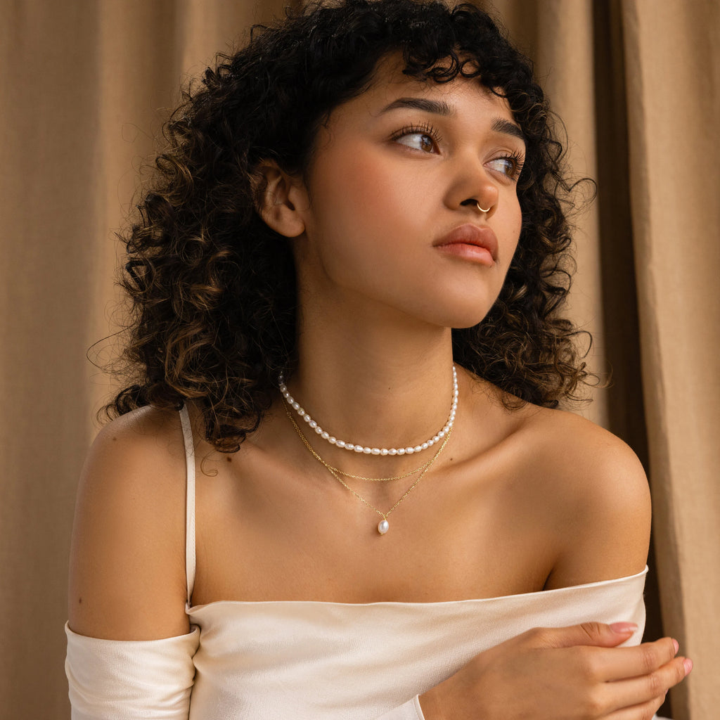 Woman with curly hair in an off-shoulder top wears the Duo Chain Pearl Necklace, featuring a pearl choker and layered chains, as she gazes to the side against a beige curtain.