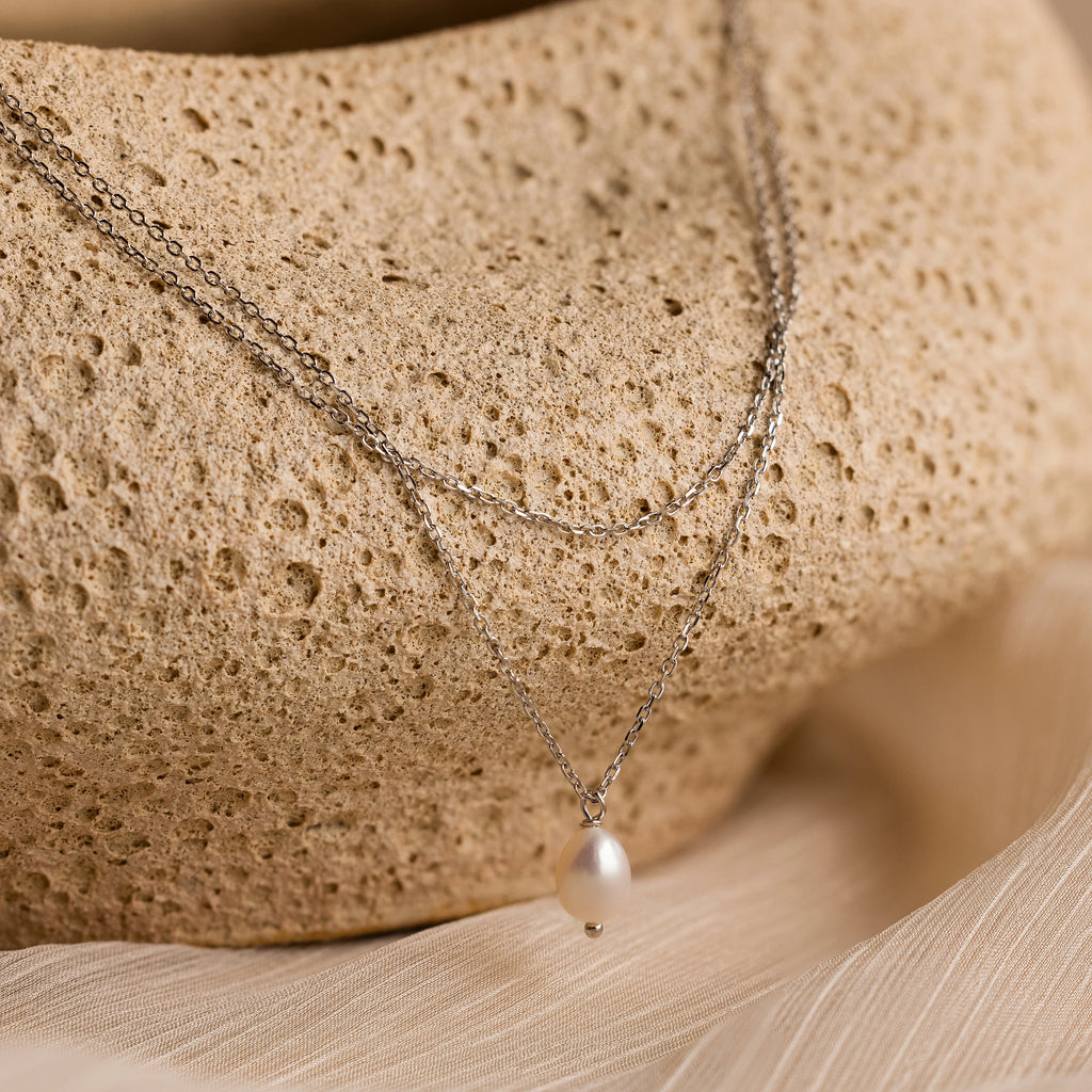 The Duo Chain Pearl Necklace features layered chains and a single pearl pendant, beautifully showcased on a textured beige stone.