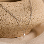 The Duo Chain Pearl Necklace features layered chains and a single pearl pendant, beautifully showcased on a textured beige stone.