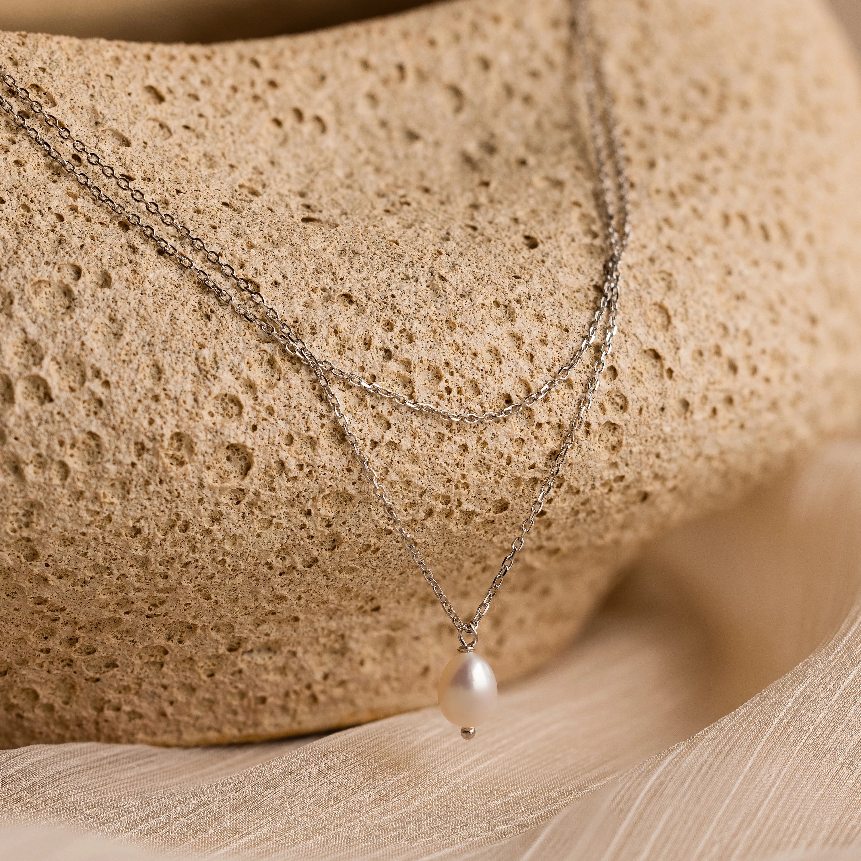 The Duo Chain Pearl Necklace features layered chains and a single pearl pendant, beautifully showcased on a textured beige stone.