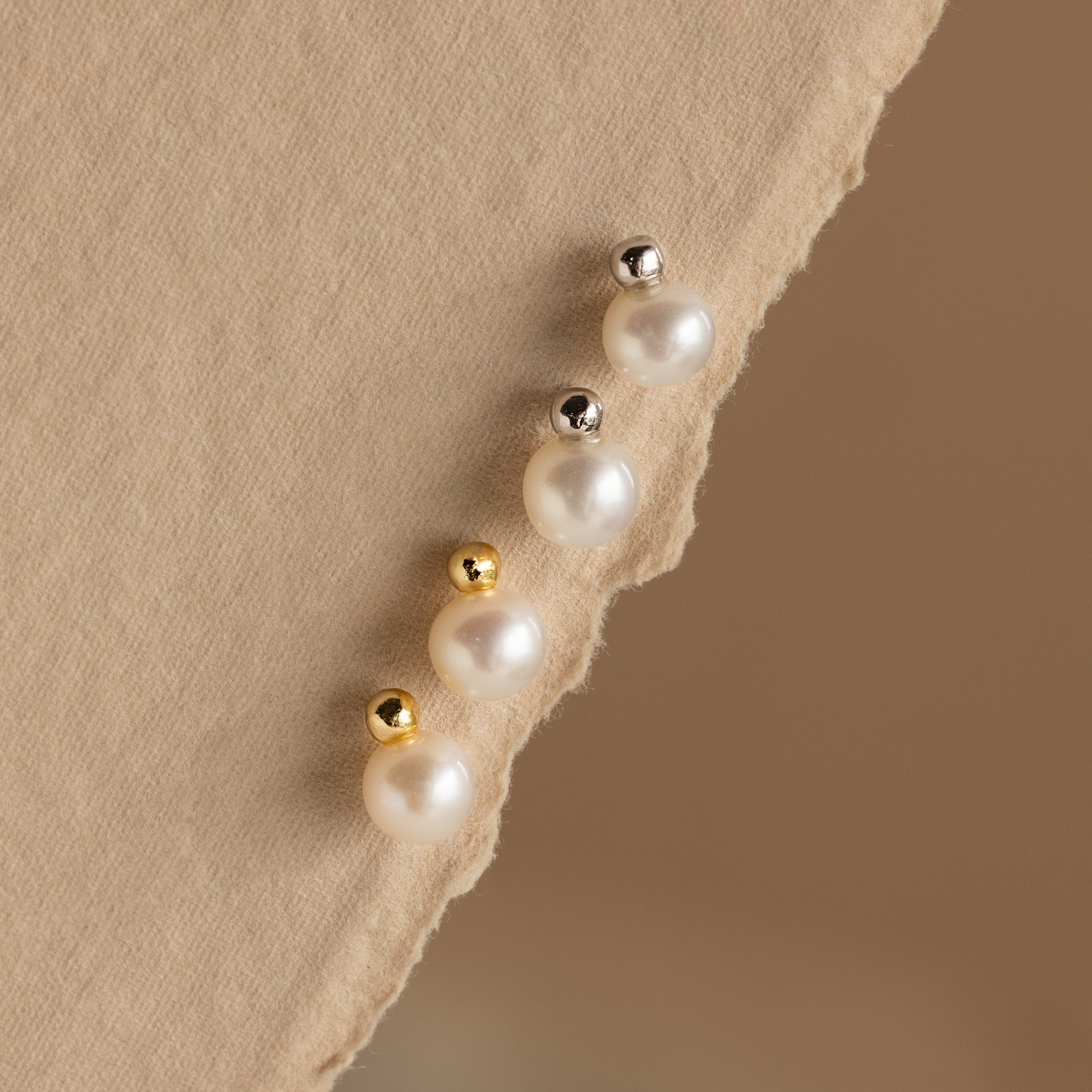 The Classic Pearl Studs are elegantly displayed on a textured beige fabric background, paired with gold and silver stud earrings for a timeless look.