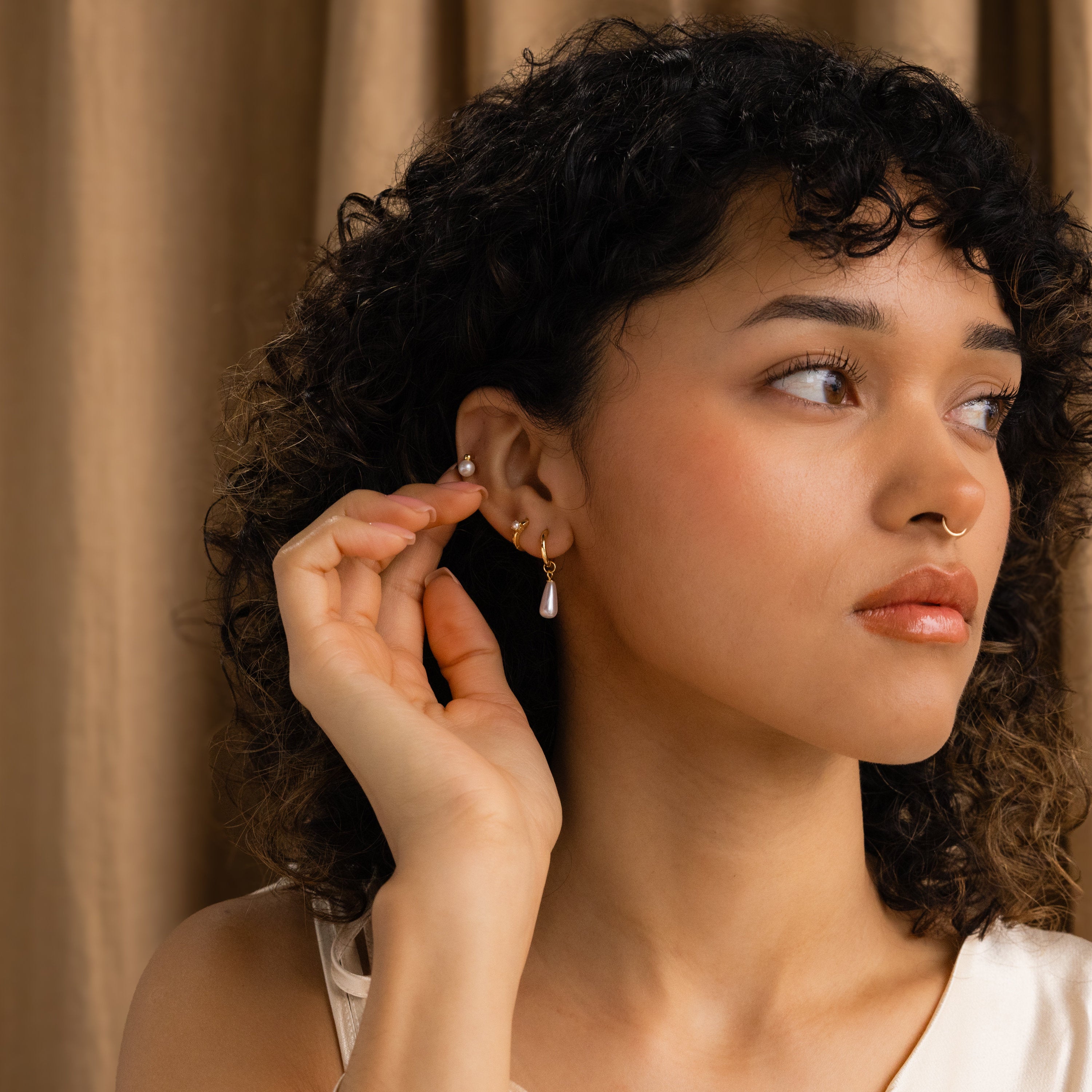 A woman with curly hair touches her Classic Pearl Studs, gazing to the side against a brown curtain backdrop.
