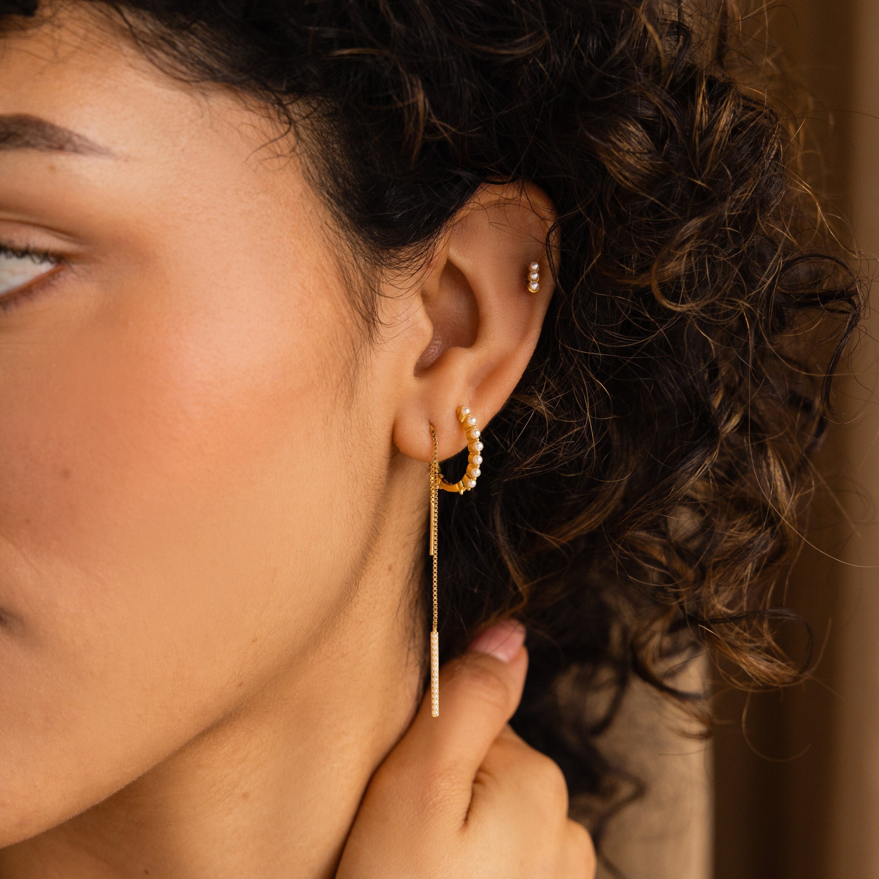 A woman with curly hair wears multiple gold earrings and holds a Pearl Bar Threader Earring, highlighting her passion for modern pearl jewelry.