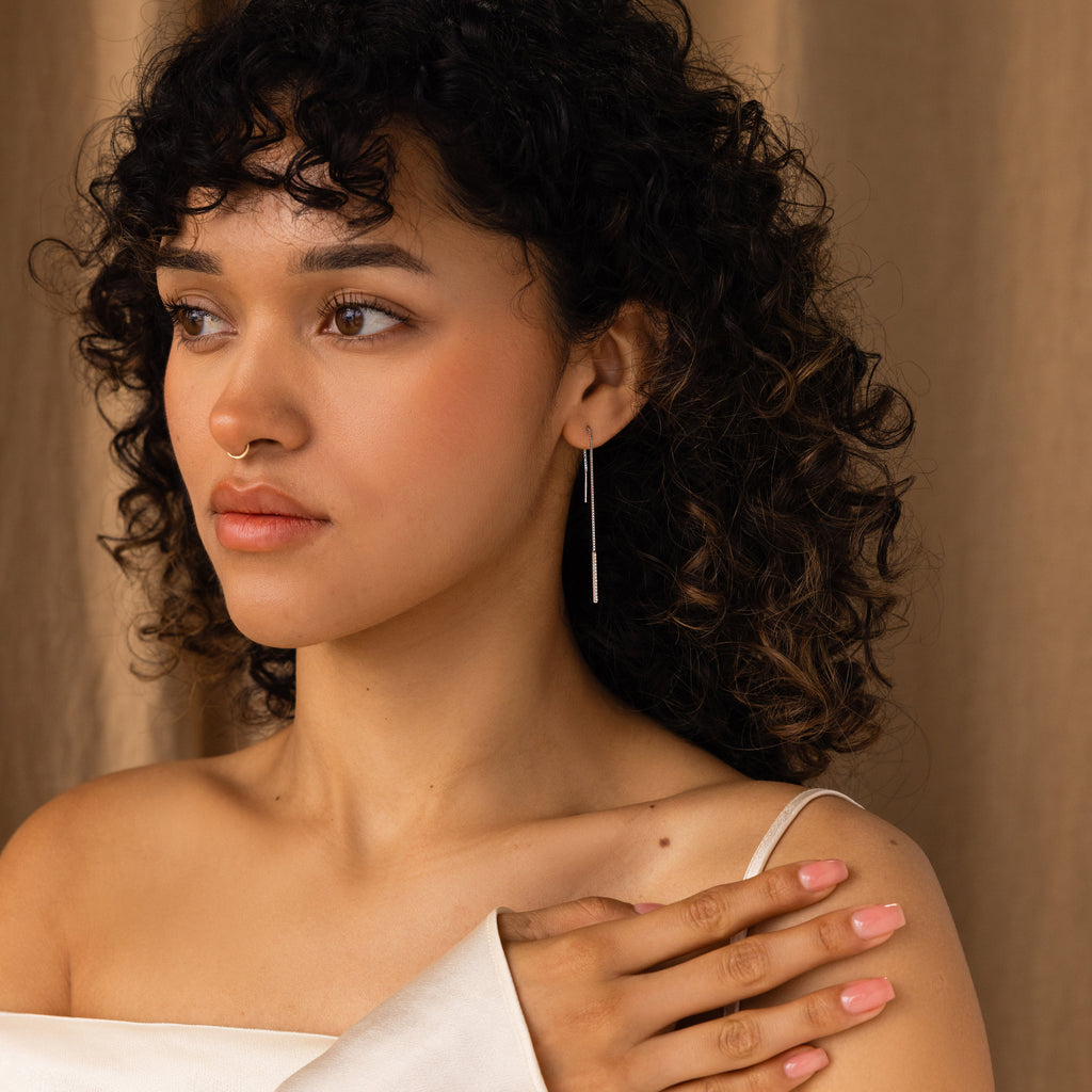 A woman with curly hair and a nose ring, in a white off-shoulder top, showcases the Pearl Bar Threader Earrings as she gently touches her shoulder.