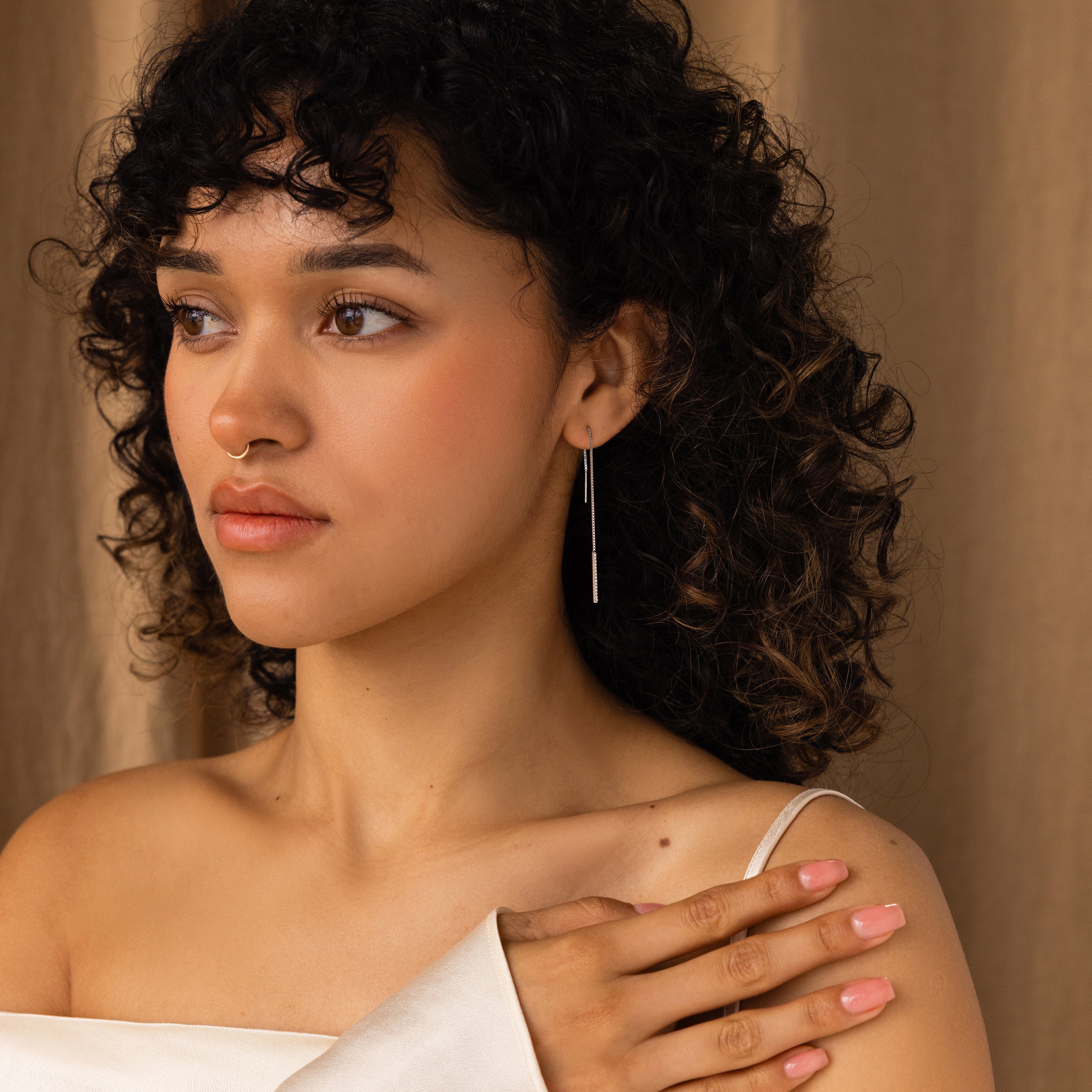 A woman with curly hair and a nose ring, in a white off-shoulder top, showcases the Pearl Bar Threader Earrings as she gently touches her shoulder.