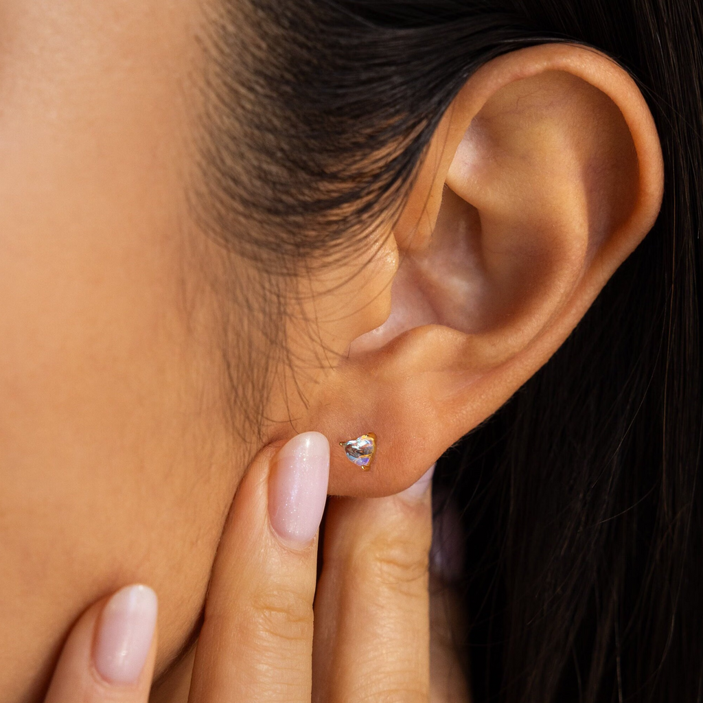 A close-up showcases a woman's ear adorned with Moonstone Heart Studs as her fingers gently touch her earlobe.