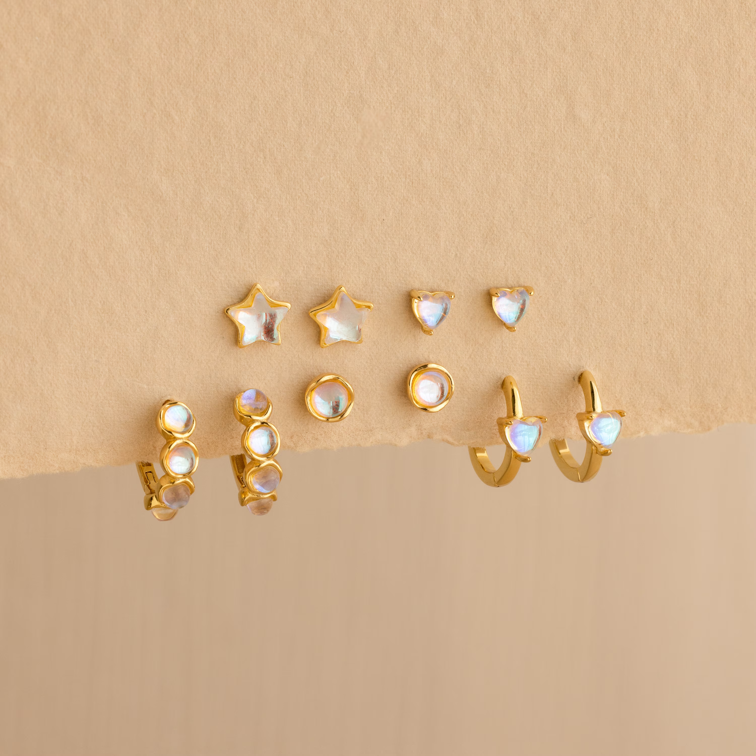 Six pairs of dainty earrings—featuring Moonstone Heart Studs plus star, round, and heart shapes—adorned with moonstone gems, displayed on a beige surface.