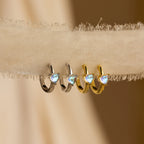 The Moonstone Heart Huggies, available in silver and gold, feature heart-shaped moonstones in a chic hoop design and are elegantly displayed on textured fabric.