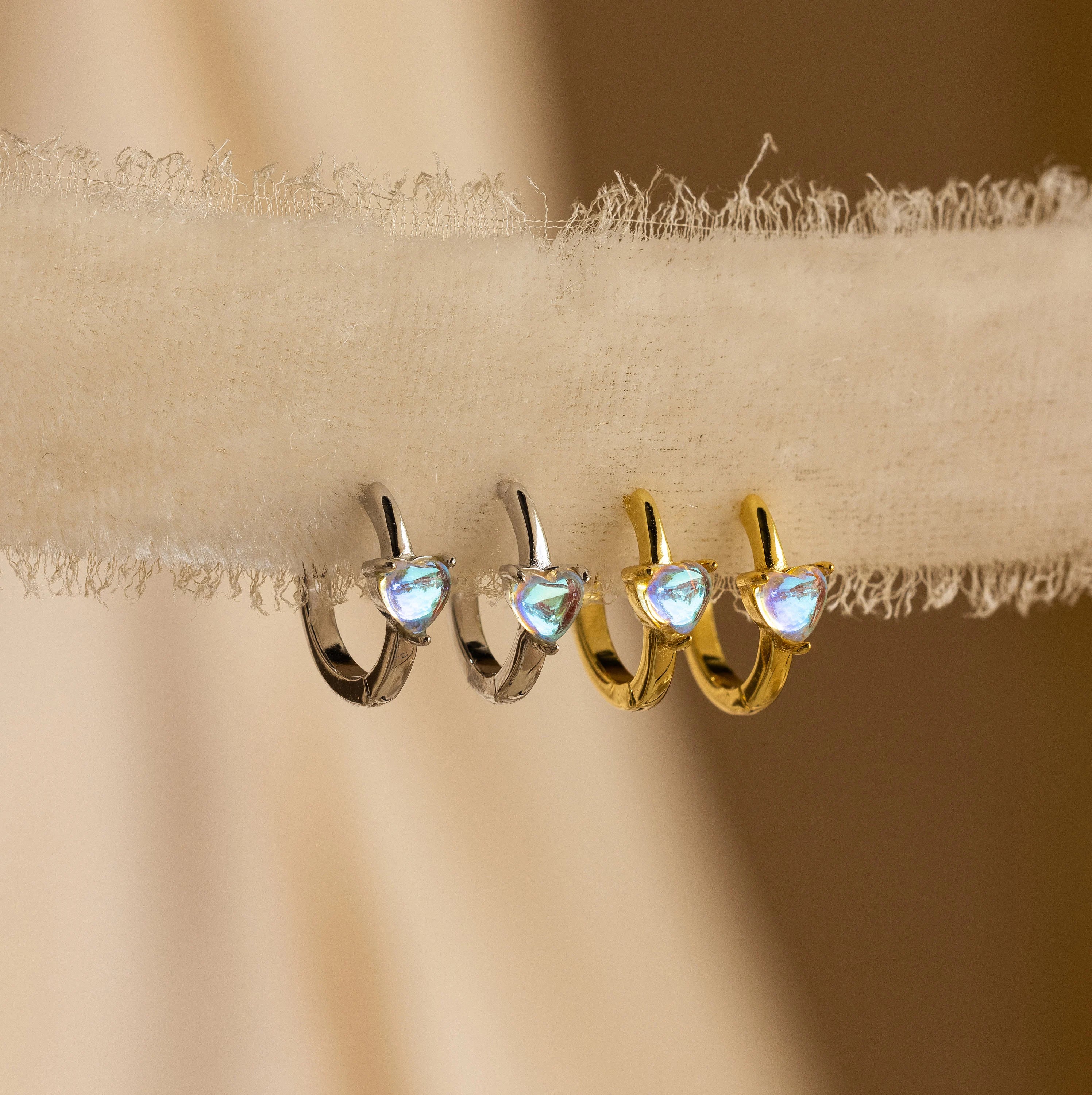 The Moonstone Heart Huggies, available in silver and gold, feature heart-shaped moonstones in a chic hoop design and are elegantly displayed on textured fabric.