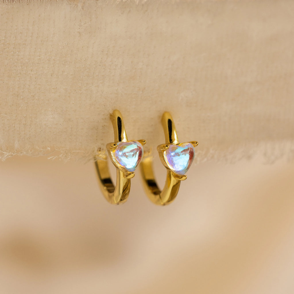 The Moonstone Heart Huggies are dainty gold hoop earrings with heart-shaped moonstones, shown against a soft beige fabric background.