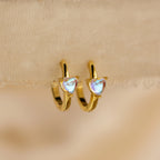 The Moonstone Heart Huggies are dainty gold hoop earrings with heart-shaped moonstones, shown against a soft beige fabric background.
