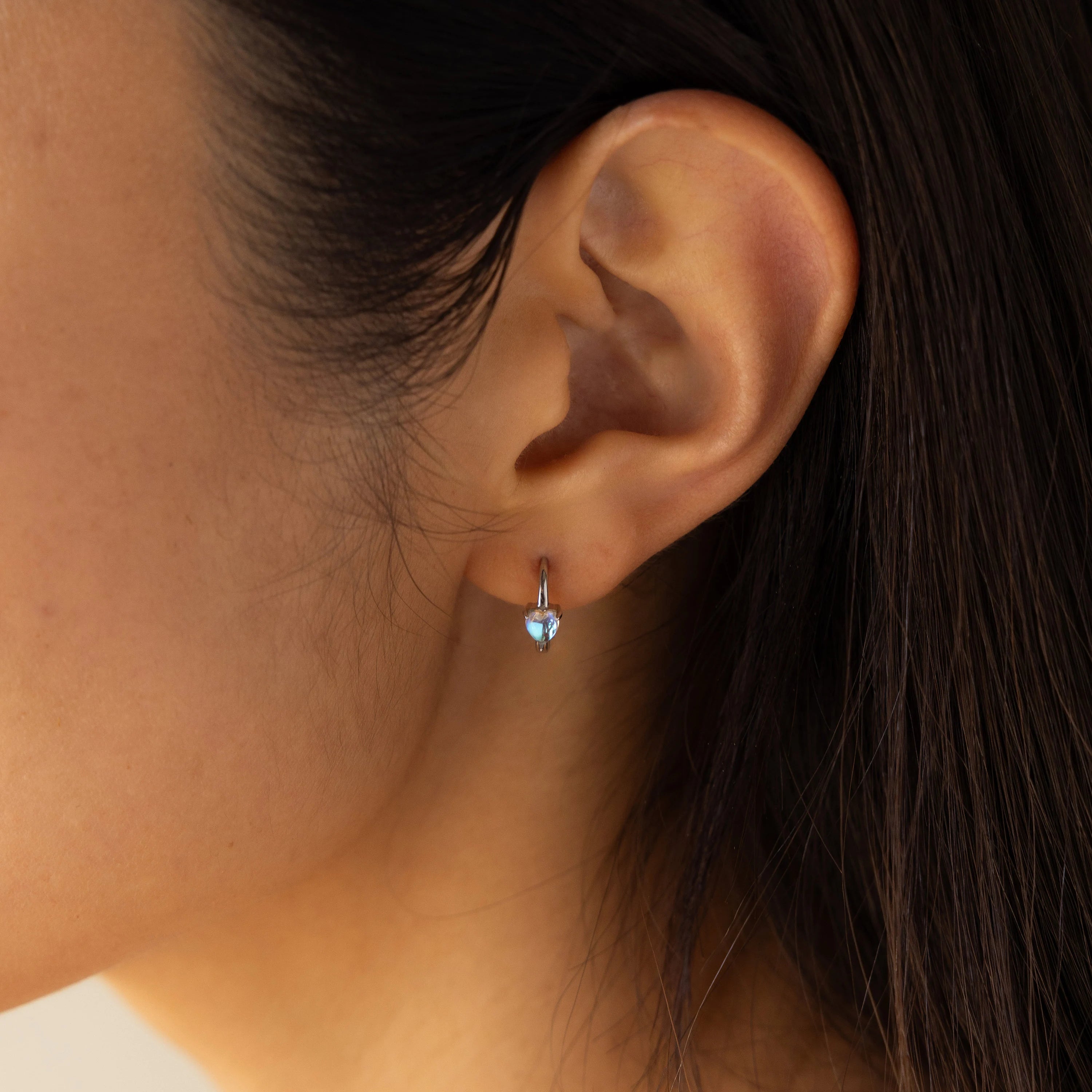 Close-up of a woman's ear wearing Moonstone Heart Huggies—delicate earrings with tiny, heart-shaped moonstones that shimmer softly in the light.