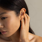 A woman with dark hair tucked behind her ear showcases the Moonstone Heart Huggies, featuring delicate heart-shaped moonstones.