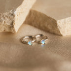 Moonstone Heart Huggies—gold hoop earrings with iridescent, heart-shaped moonstones—rest on a beige textured surface near stone slabs. These dainty earrings are perfect for any occasion.