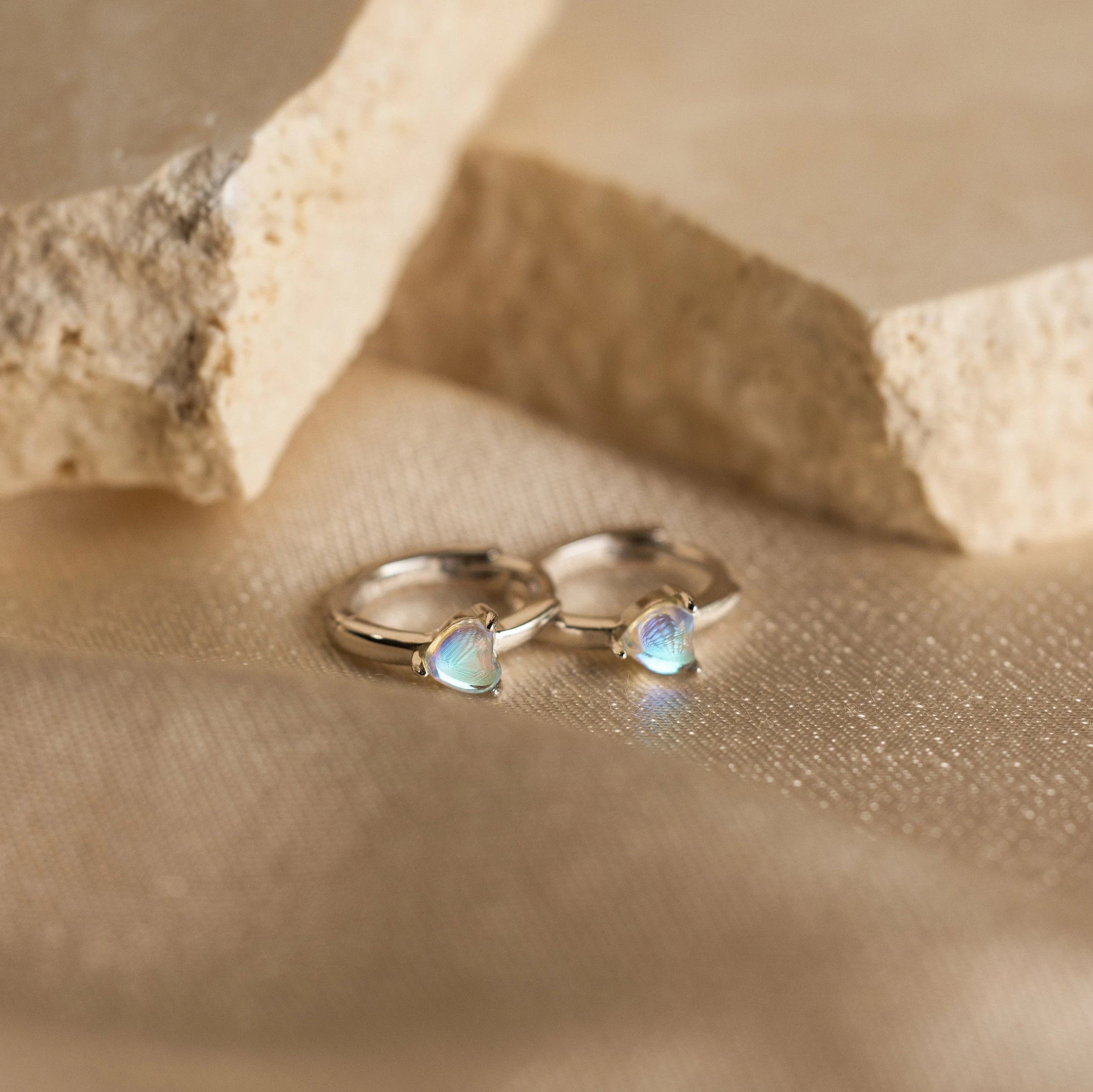 Moonstone Heart Huggies—gold hoop earrings with iridescent, heart-shaped moonstones—rest on a beige textured surface near stone slabs. These dainty earrings are perfect for any occasion.