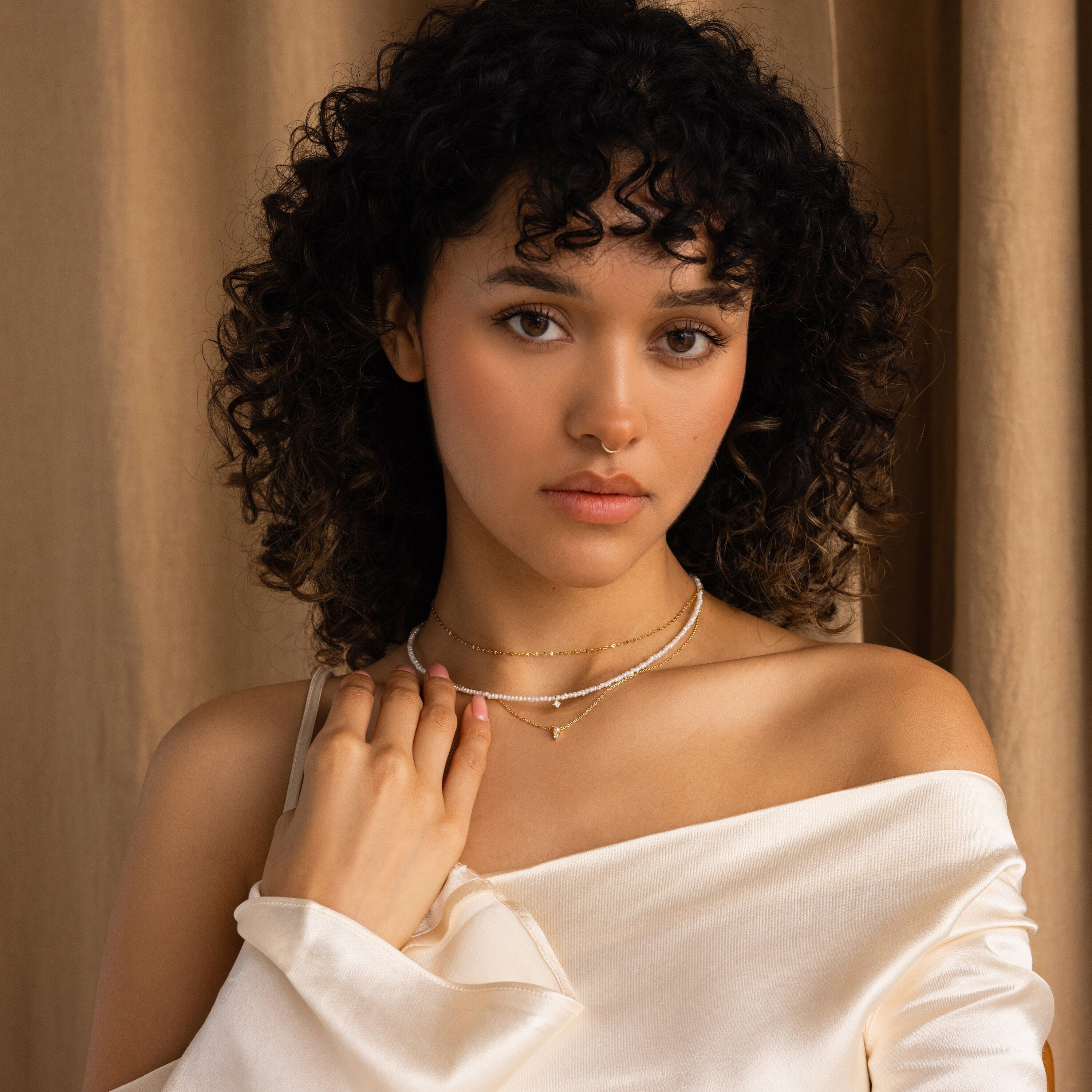 Woman with curly hair, in an off-shoulder white top, stands against a beige curtain wearing the Pearl & Diamond Duo Necklace.