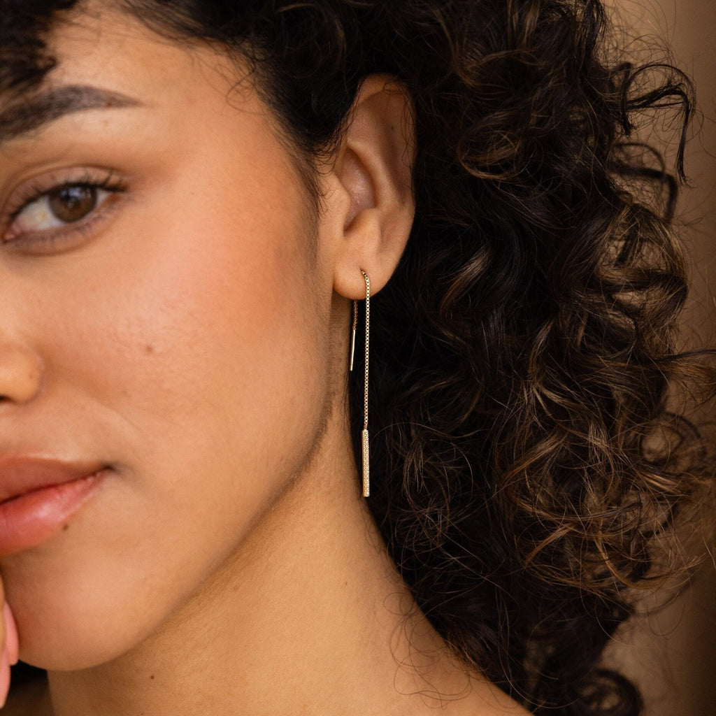 A woman with curly hair rests her chin on her hand, wearing delicate Pearl Bar Threader Earrings.