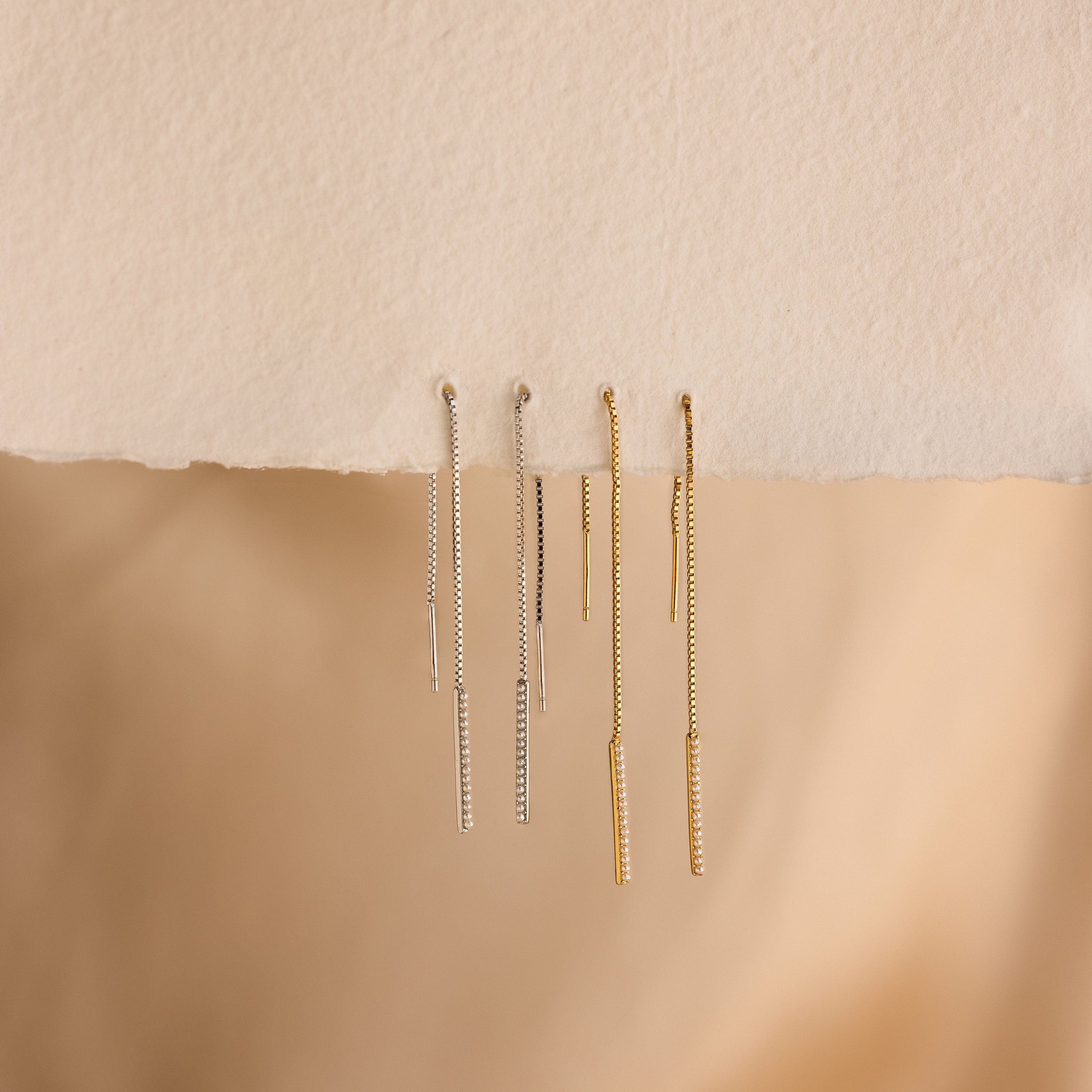 Four pairs of dainty earrings, including the Pearl Bar Threader Earrings, are displayed on a textured beige surface.