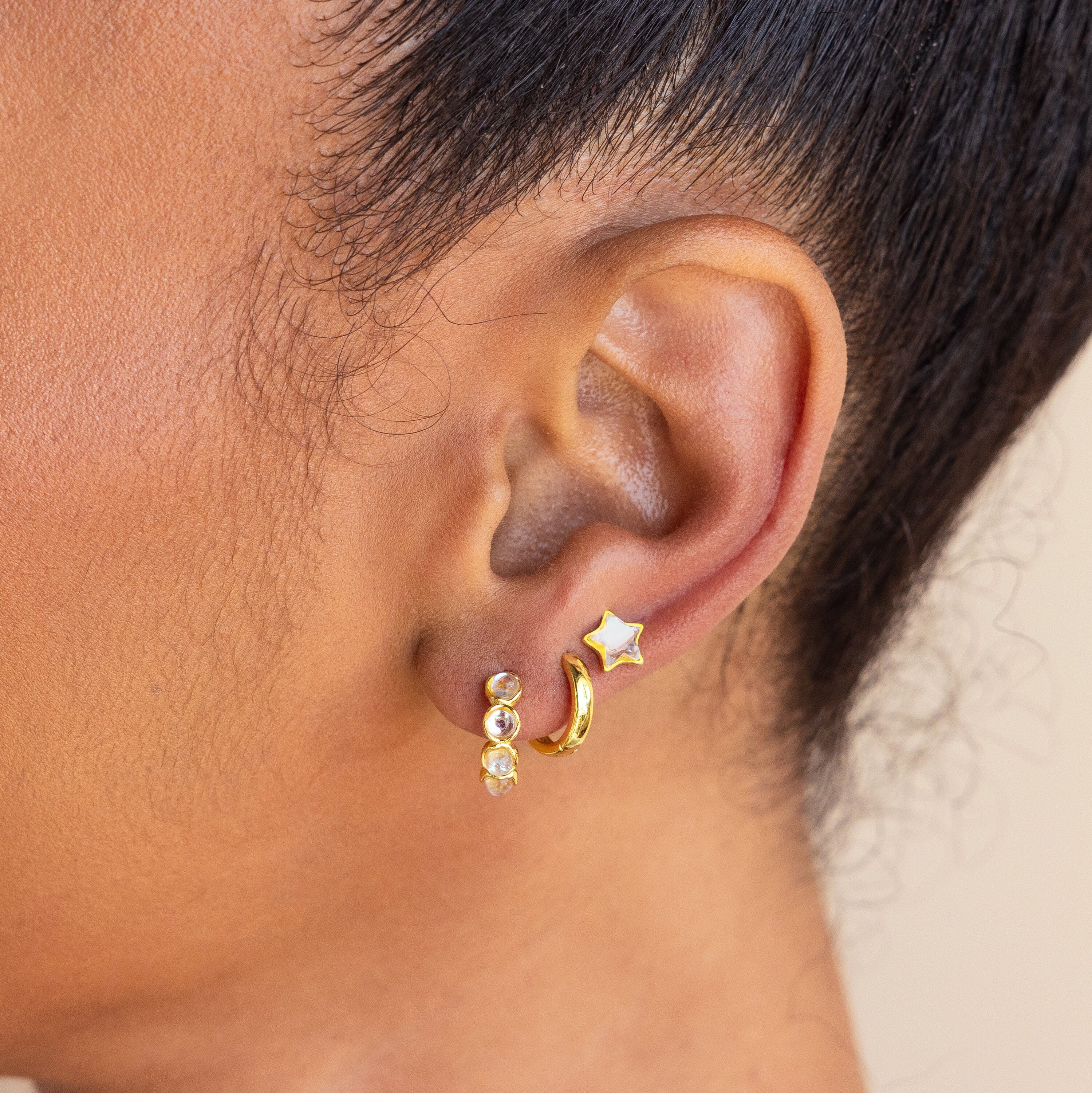 Close-up of an ear wearing celestial jewelry: Moonstone Star Studs, a gold hoop, and a small hoop with three gems.