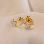 Moonstone Star Studs with gold star-shaped design and iridescent centers shimmer on a soft beige fabric background.